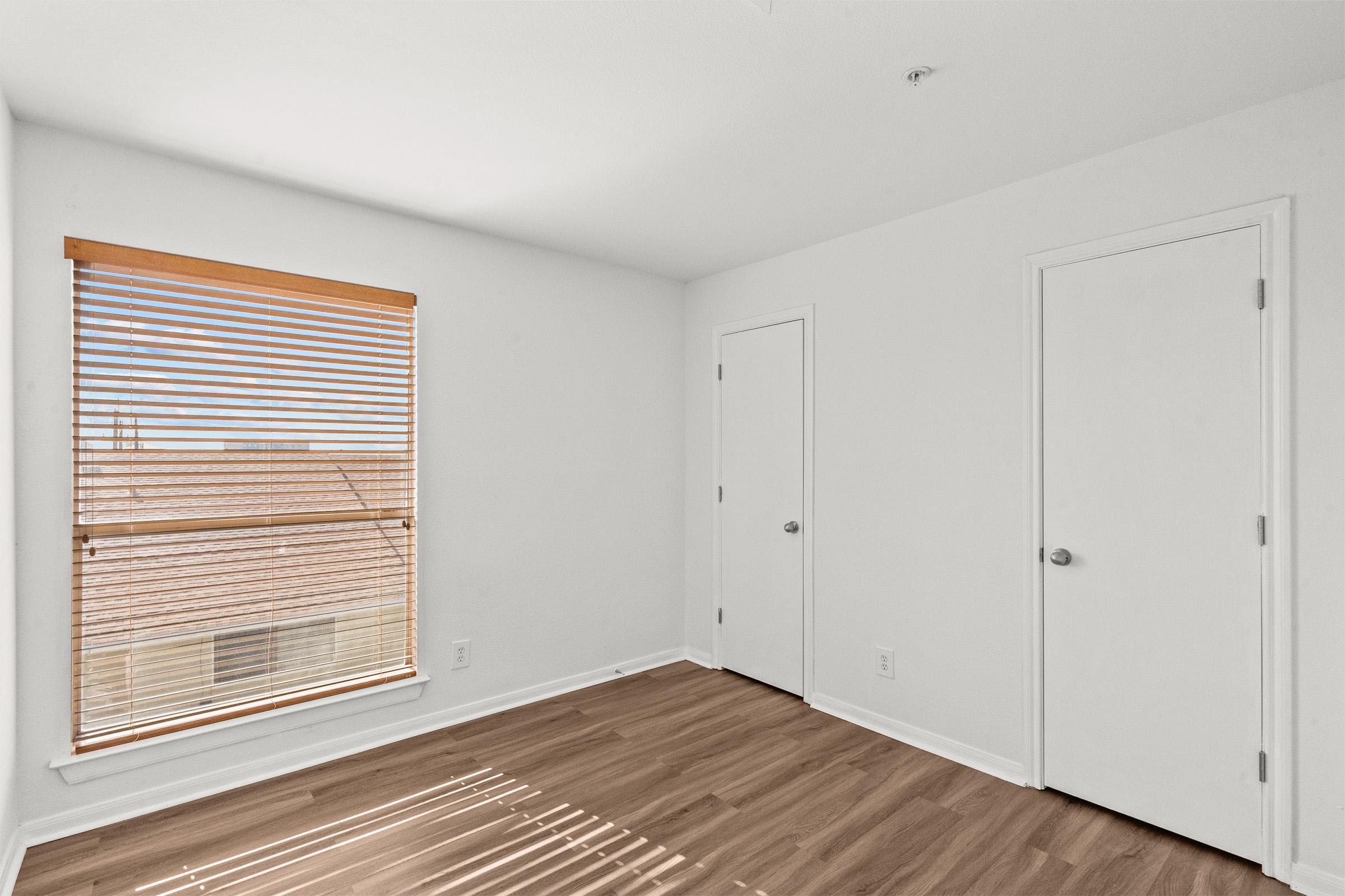 Empty room featuring light-colored walls and laminate flooring. A window with wooden blinds allows natural light to enter, casting shadows on the floor. There are two closed doors on the right side of the image, adding to the room's simplicity and minimalism.
