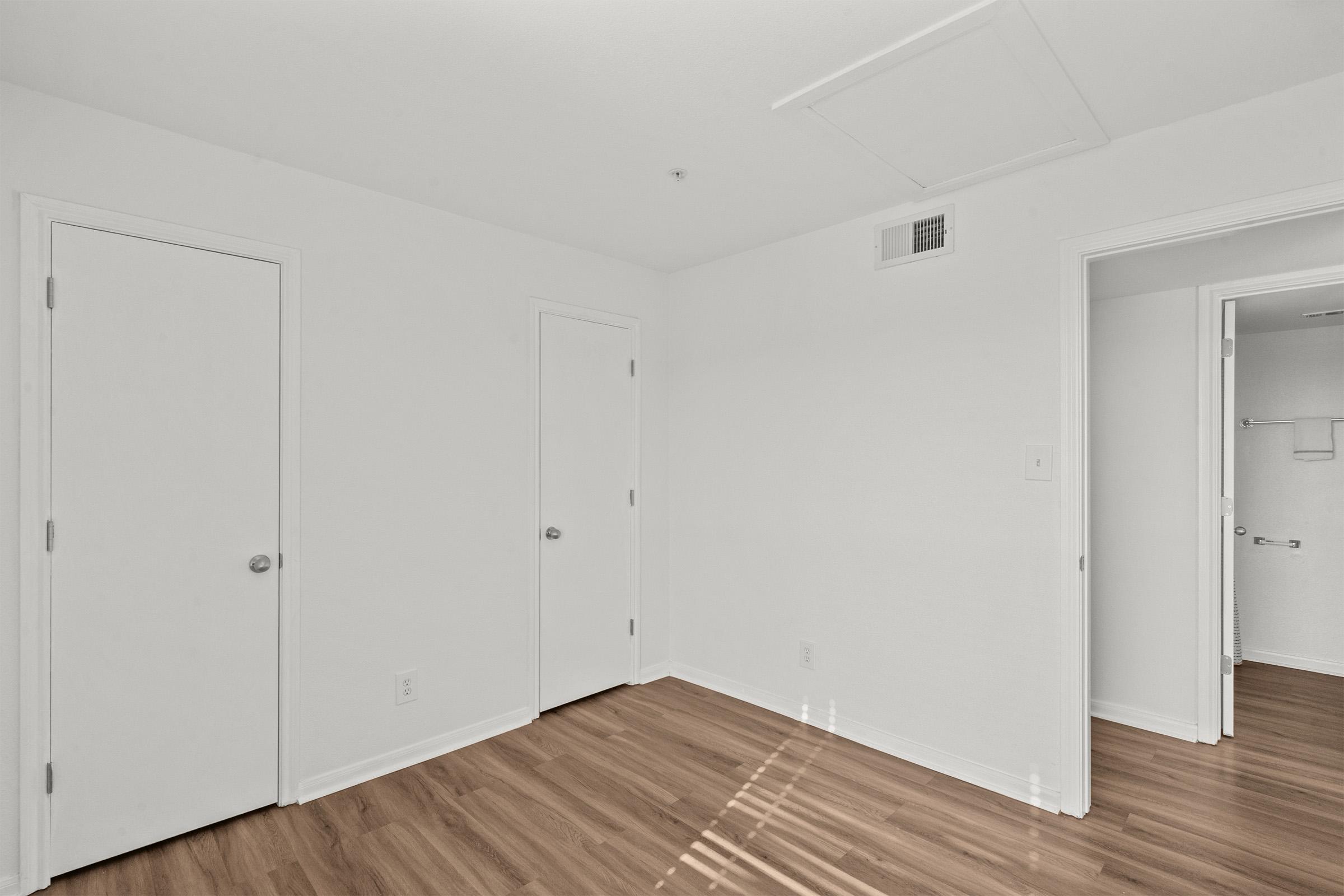 A bright, empty room with light-colored walls and wooden flooring. Two closed doors are visible on the left side, and an additional doorway leads to another space on the right. Natural light casts soft shadows on the floor, creating a clean and spacious atmosphere.