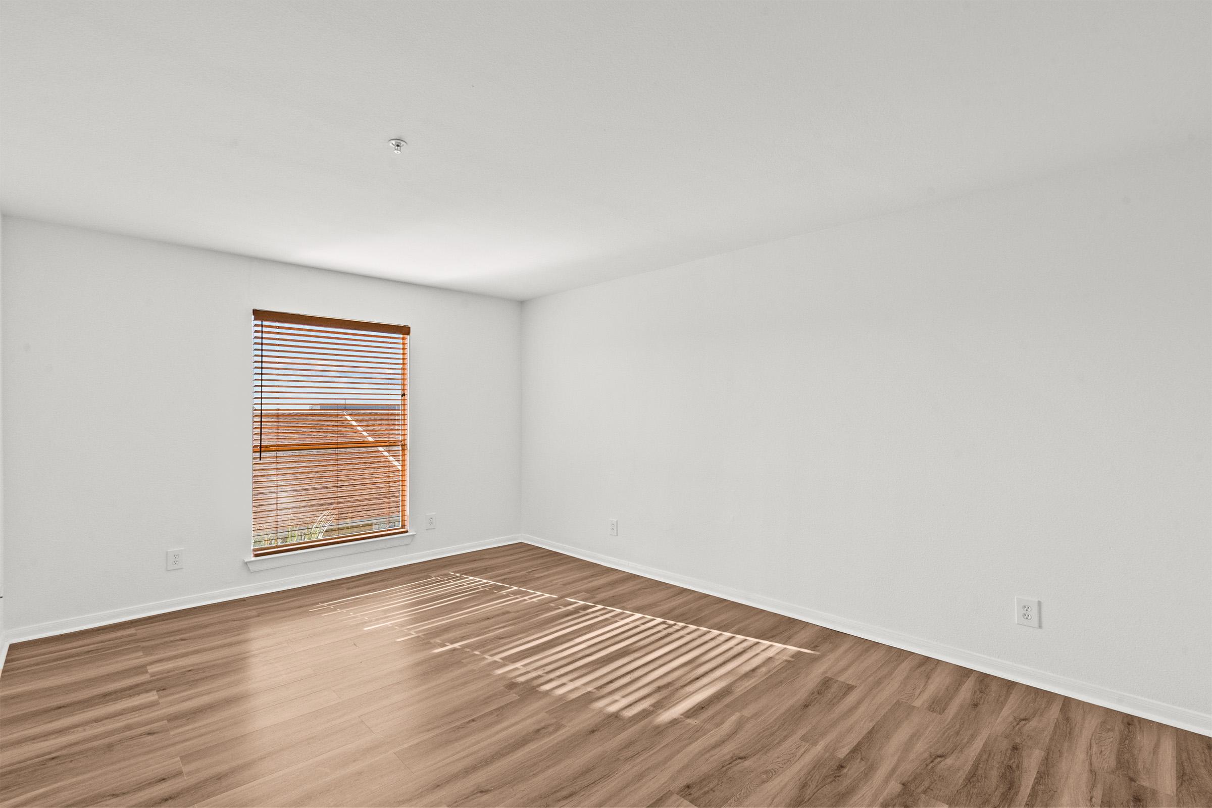 A spacious, empty room with light-colored walls and wooden flooring. Natural light streams in through a window with blinds, casting shadows on the floor. The space is devoid of furniture, creating a clean and minimalistic atmosphere.