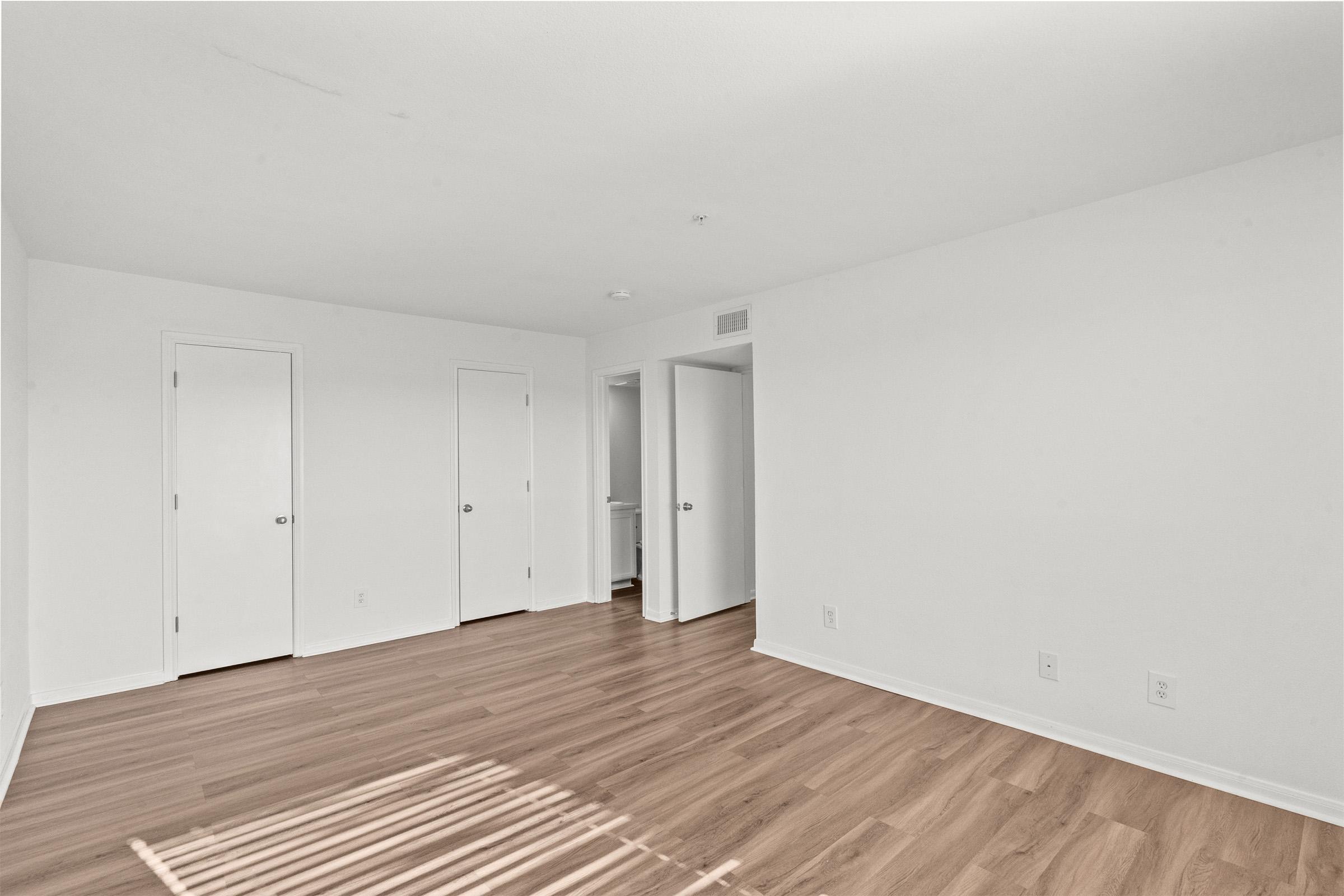 A bright, empty room with light-colored walls and wooden flooring. Two doors are visible on the left side, and a third door is on the right, leading to another space. Natural light creates subtle shadows on the floor, emphasizing the room's spaciousness and clean aesthetic.