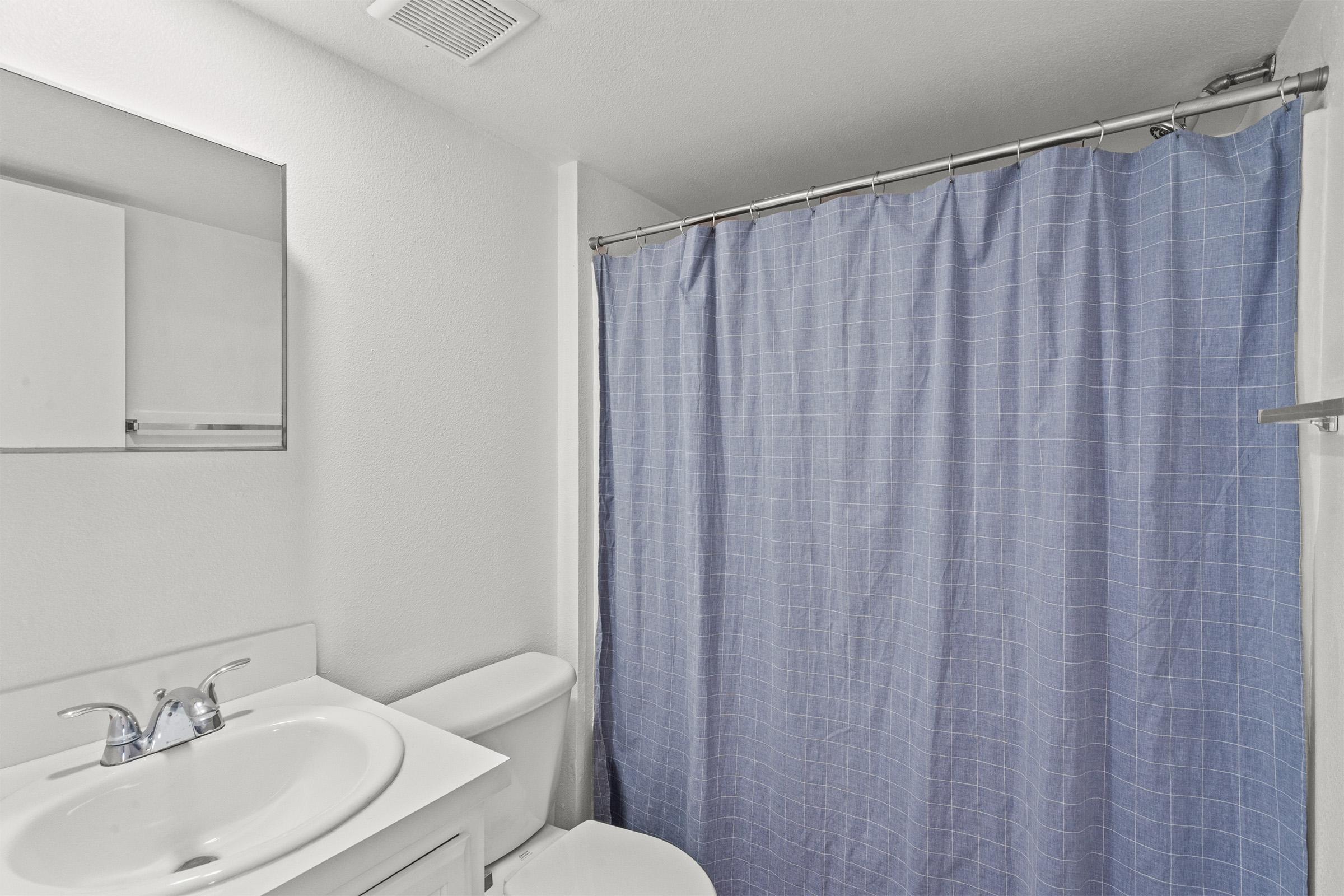 A clean bathroom with a white sink and toilet. There is a large mirror above the sink and a light blue shower curtain at the side, providing a simple and tidy appearance. The walls are painted white, giving a bright and airy feeling to the space.
