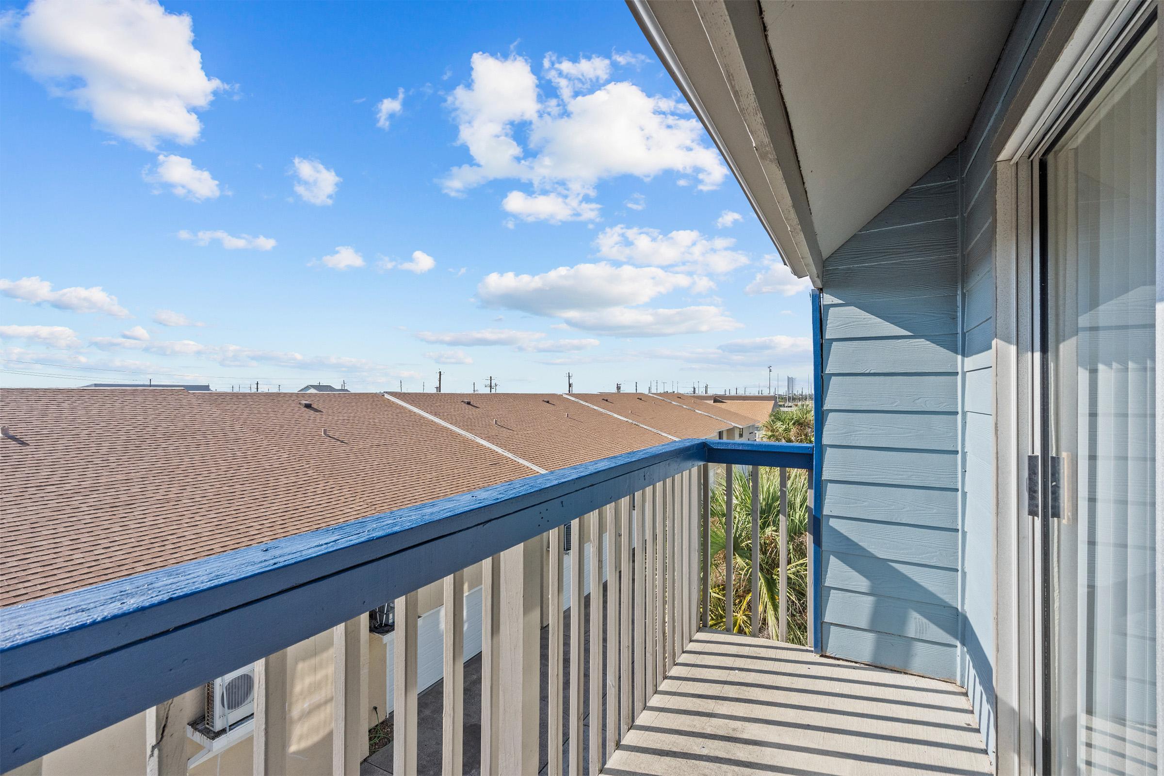 View from a balcony overlooking rooftops under a bright blue sky with scattered clouds. The balcony has a blue railing and is part of a building with light-colored siding. In the distance, utility poles are visible against the skyline.