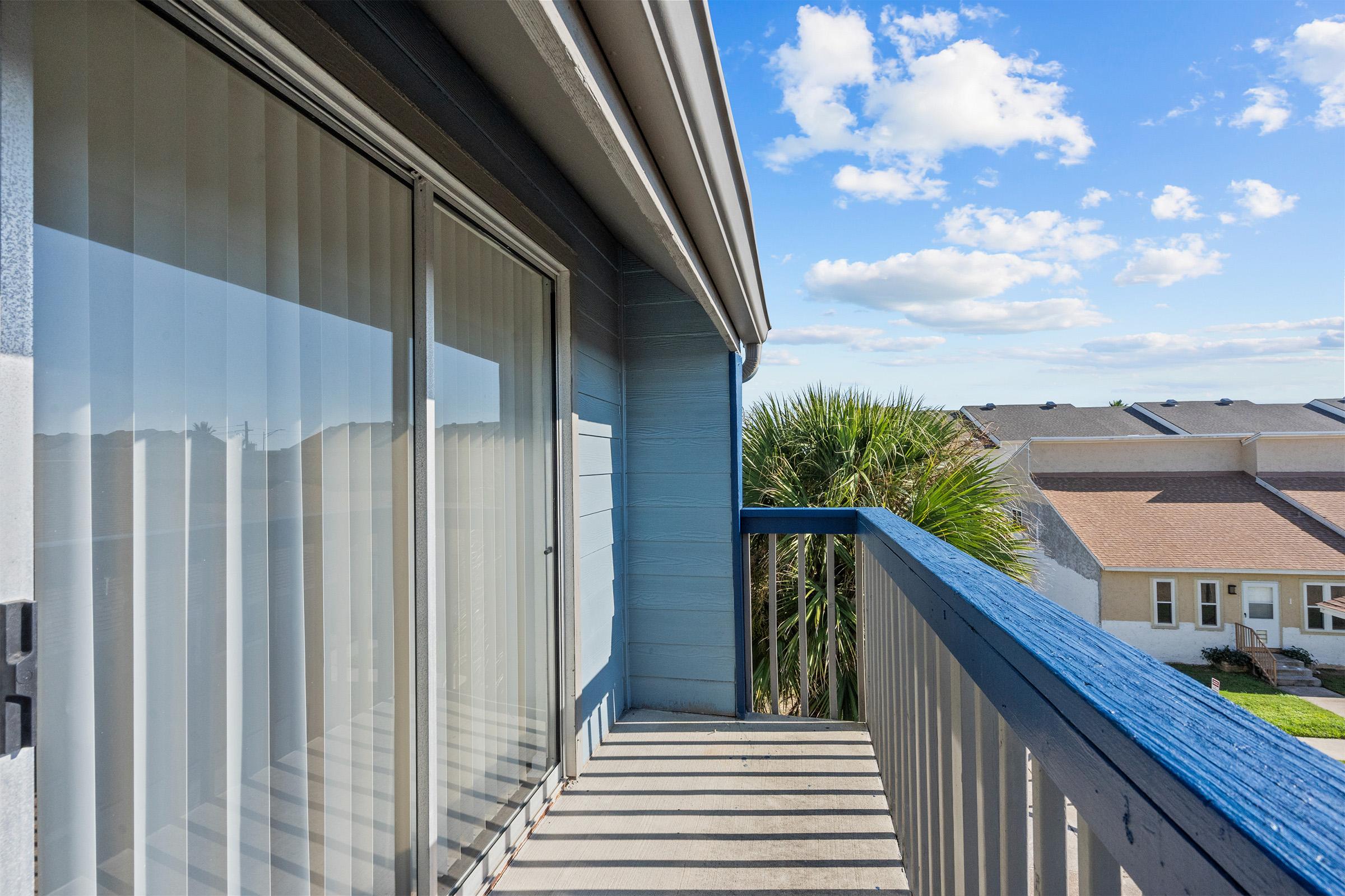 A balcony view featuring sliding glass doors, blue railings, and a palm tree in the background. The sky is partly cloudy, and there are residential buildings in the distance. Sunlight casts shadows on the balcony floor.