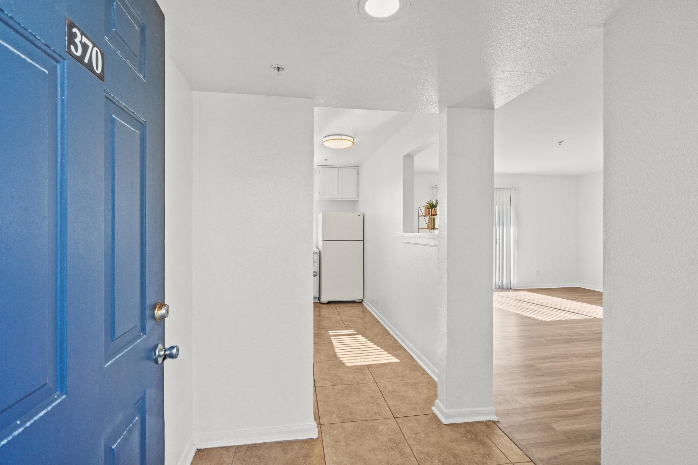 A brightly lit hallway leading to a living area, featuring a blue door labeled "370." On the left, there is a glimpse of a kitchen with a white refrigerator. The floor is a mix of tile and laminate, and natural light streams in from a window in the adjacent room.