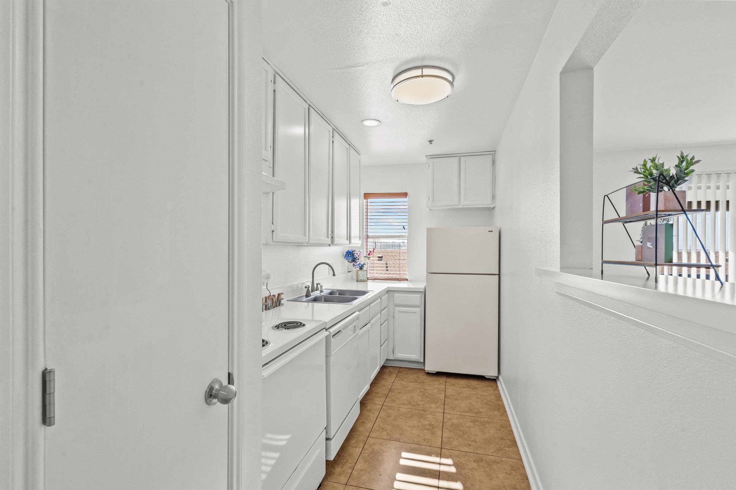 Bright and airy kitchen with white cabinetry, a refrigerator, and a sink. The room features tile flooring and a window with blinds that lets in natural light. The layout includes a space for cooking and a small area for dining.