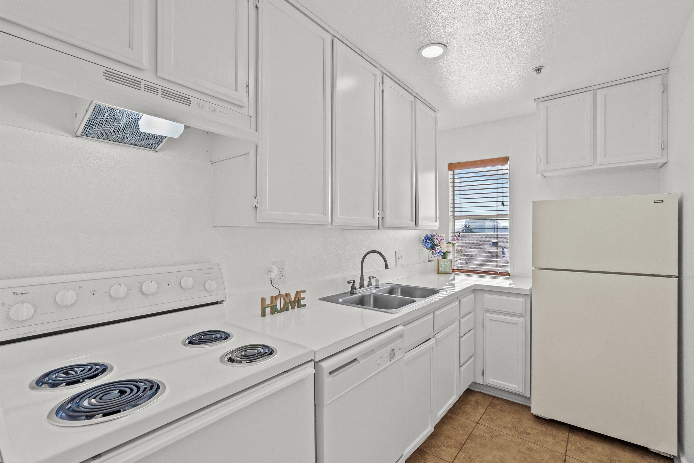 A bright, modern kitchen featuring white cabinets and appliances, including a stove, dishwasher, and refrigerator. The countertop is clean and uncluttered, with a small decorative sign reading "HOME." A window allows natural light to filter in, illuminating the space. The floor is tiled in a neutral color.