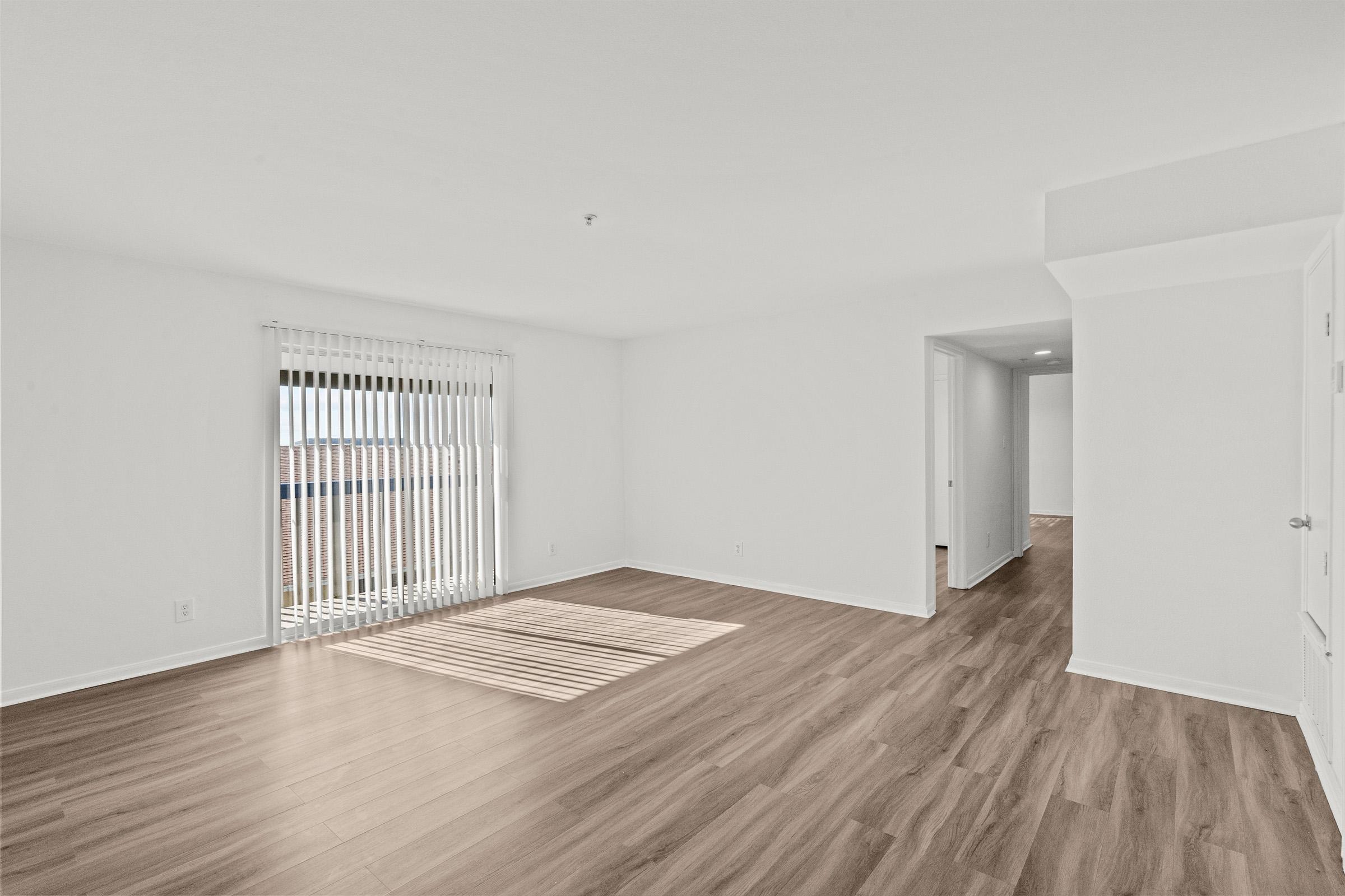 Bright and spacious interior of a living room with light wood flooring and white walls. Large window with vertical blinds allows natural light to enter. Empty space, with a glimpse of a doorway leading to another room, creating a clean and modern atmosphere.