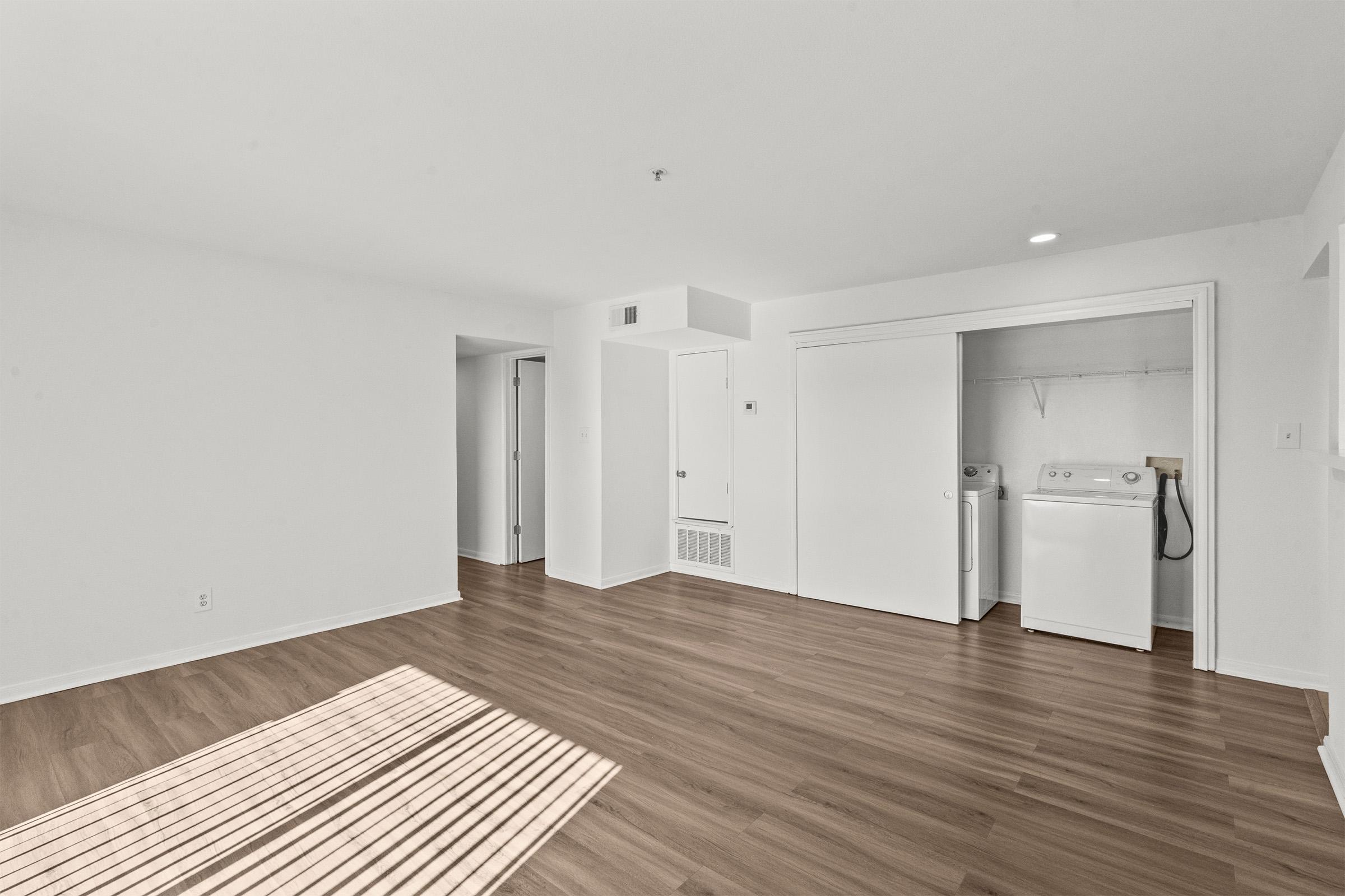 Empty room with light-colored walls and wooden flooring. There's a closet with white doors on the left, and a laundry area with a washing machine in the back corner. Natural light casts a shadow of blinds on the floor, creating a bright and spacious atmosphere.