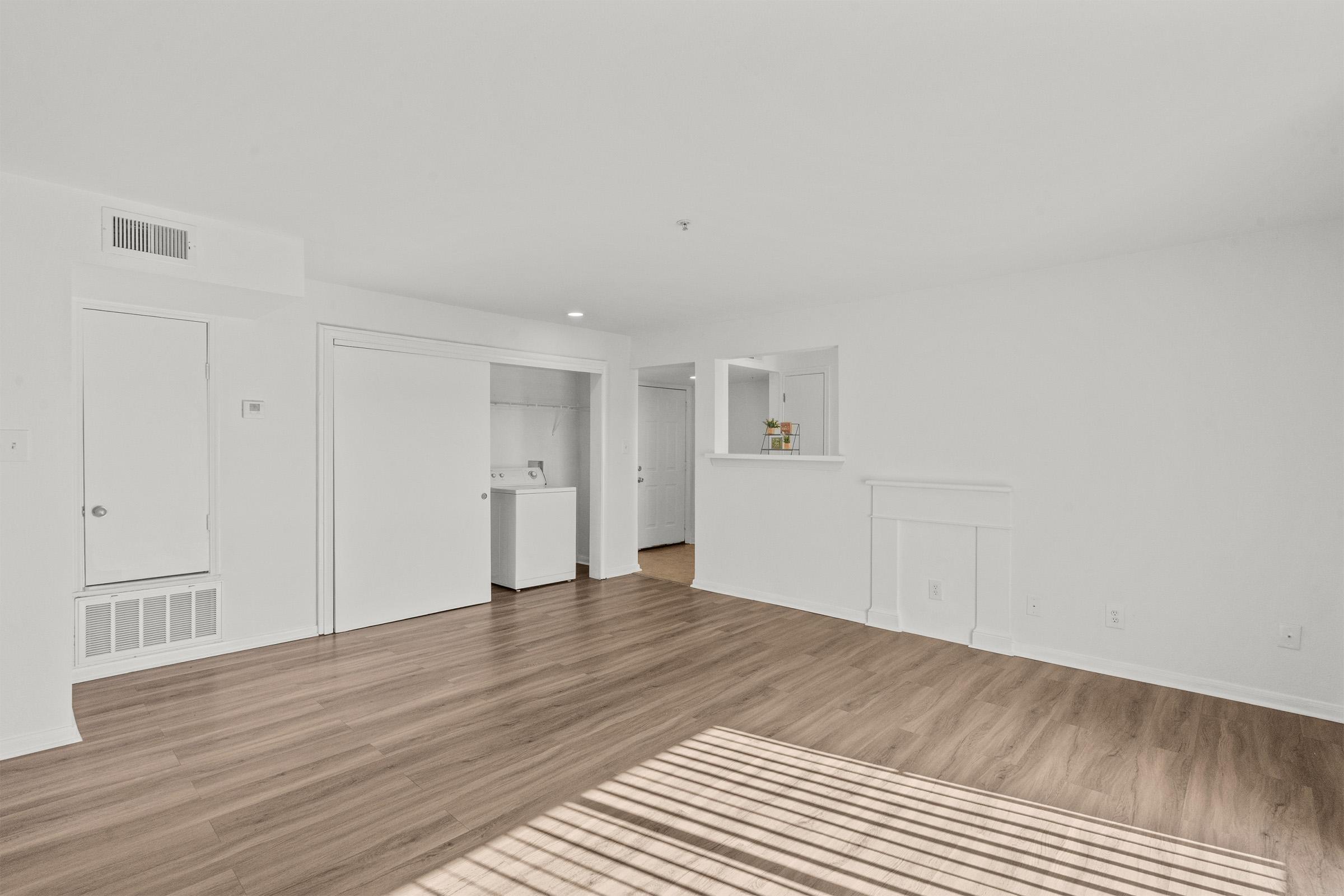 A bright, spacious living area featuring light-colored walls and wood-like flooring. There are two closed doors, one leading to a laundry space, and another that may lead to a closet or bathroom. A small opening connects to a kitchenette. Soft shadows from window blinds create a warm ambiance.