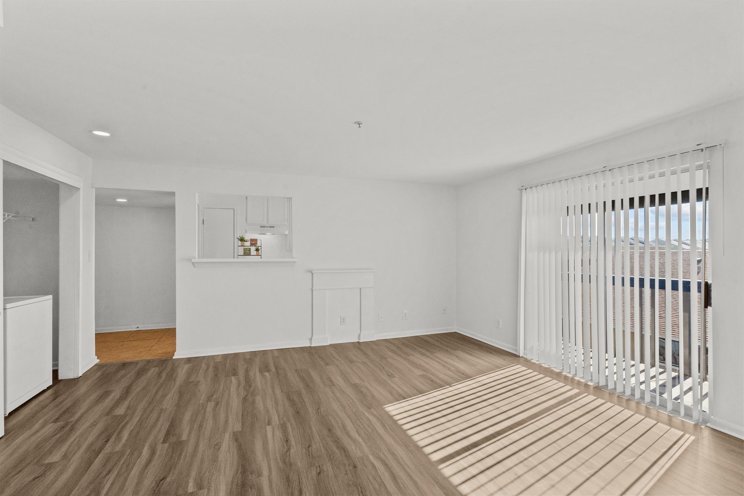 Bright and airy living space featuring light wood flooring, large windows with blinds, and a white wall color. There is a small kitchen area visible in the background and a hallway leading to another room. Sunlight casts shadows on the floor, creating a warm atmosphere.