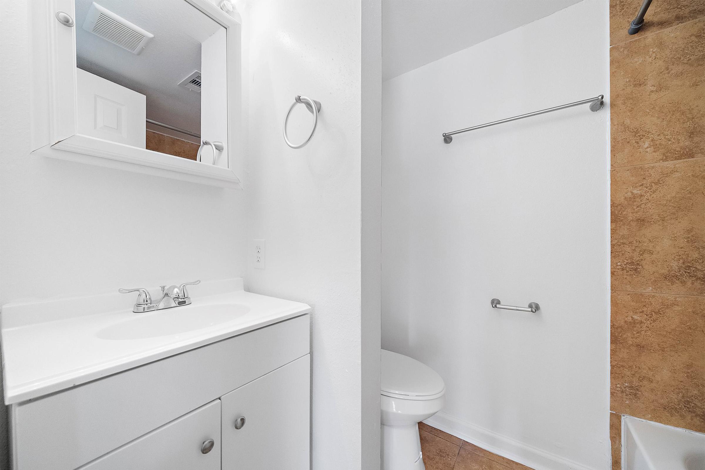 A clean and modern bathroom featuring a white vanity with a sink, a mirror above, a toilet, and a shower area. The walls are painted white, and one side has beige tiles. A towel bar is visible on the wall, adding to the functionality of the space. The overall design is simple and uncluttered.