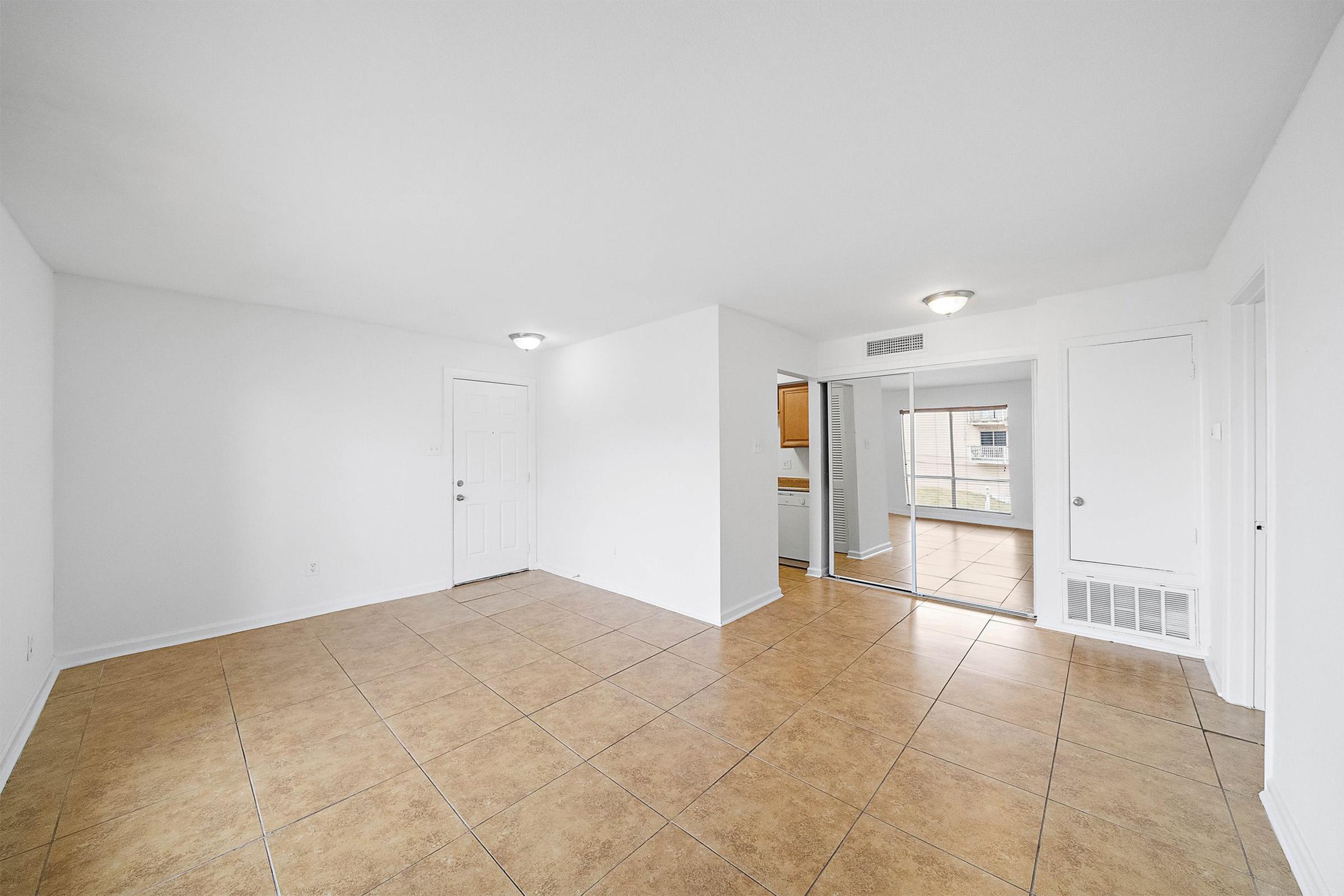 Spacious, empty living area with tiled floor and white walls. Includes a doorway leading to a kitchen area and a sliding glass door to a patio or balcony. Bright lighting from ceiling fixtures enhances the openness of the space. Perfect for customization and interior design.