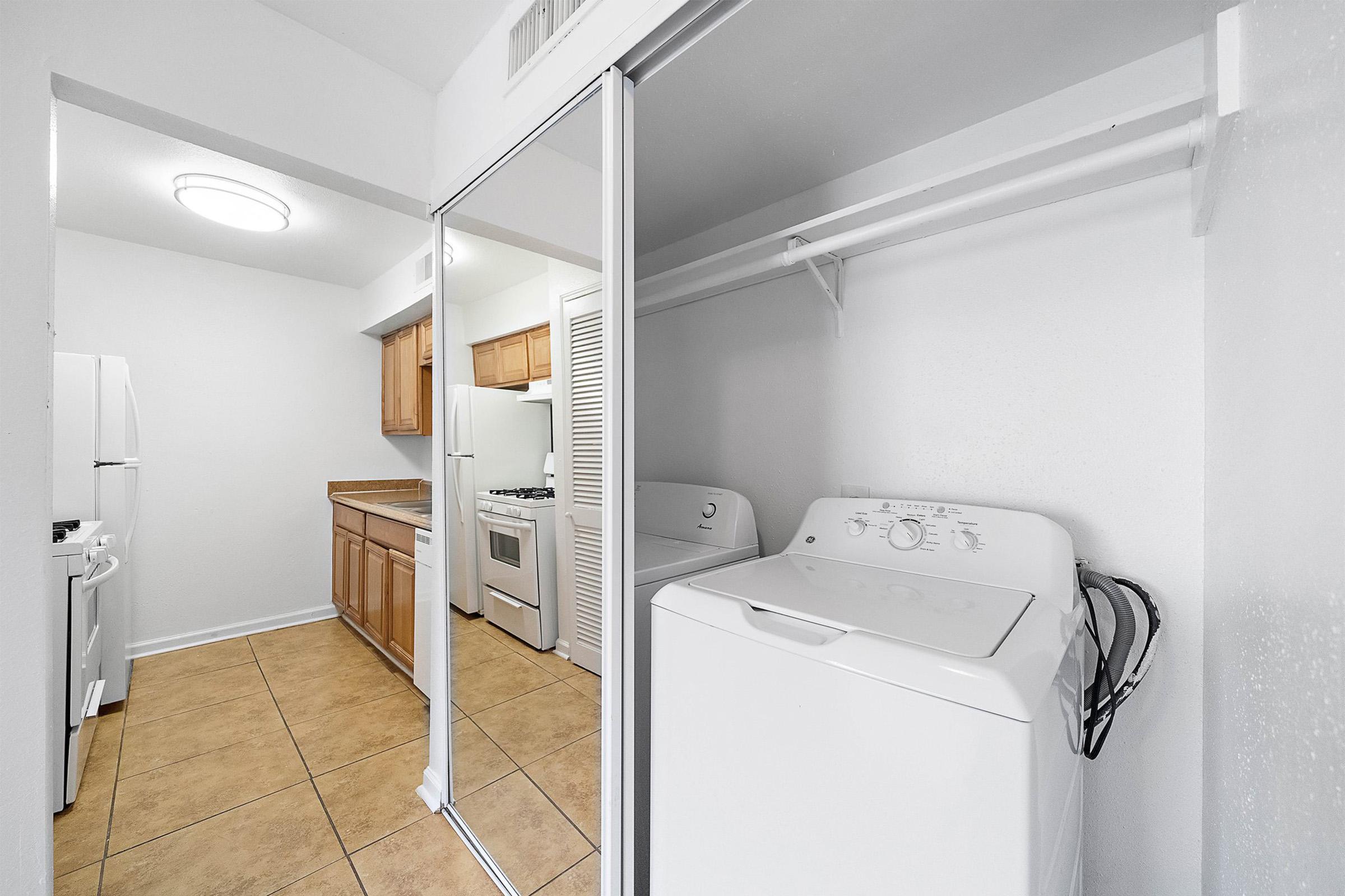 A clean, modern laundry area featuring a top-load washing machine next to a mirrored closet. The surrounding kitchen space includes wooden cabinets, a gas stove, a refrigerator, and tile flooring, creating a bright and functional household environment.