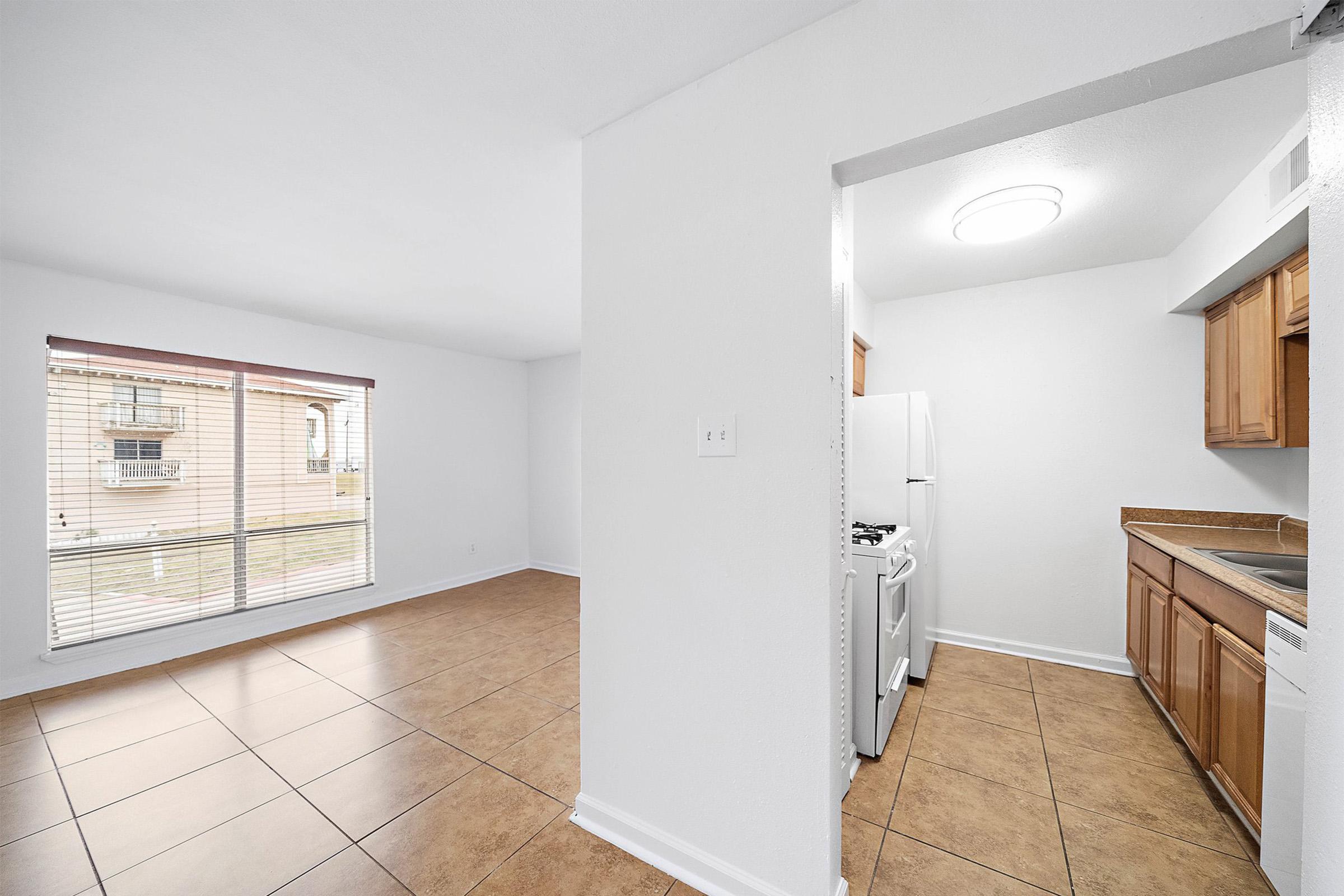 A spacious living area with tile flooring, featuring a large window that lets in natural light. To the right, there's a kitchen with wooden cabinetry and a white gas stove. The walls are painted in a light color, creating a bright and inviting atmosphere.