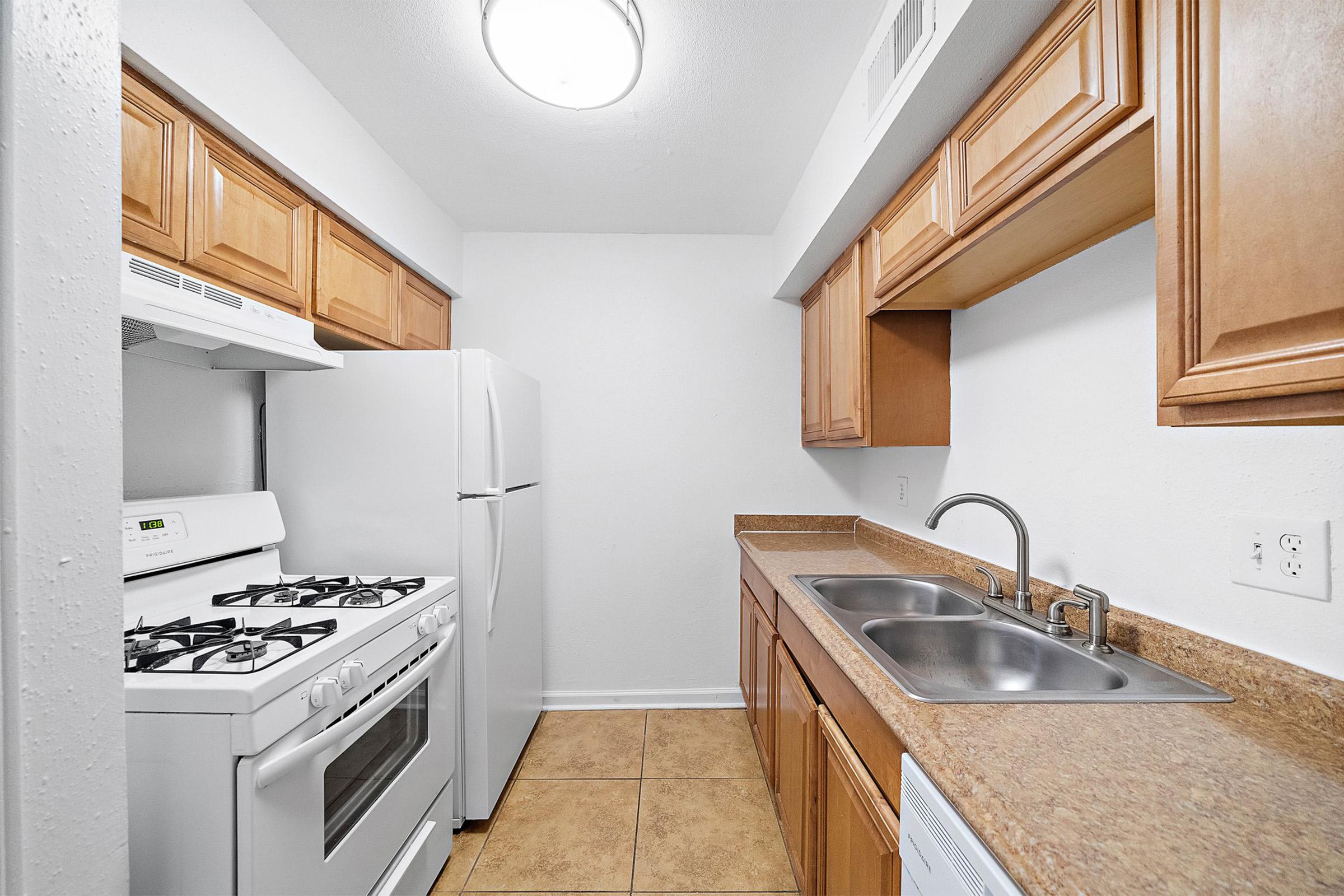 A modern kitchen featuring light wood cabinets, a white refrigerator, a gas stove with an oven, and a double sink. The countertops are made of a tan material, and there is tile flooring. The walls are painted white, and there is ceiling lighting for illumination.