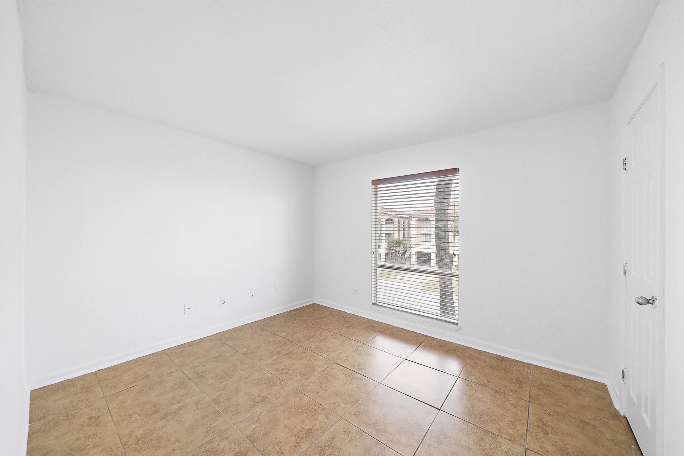 Empty room with white walls and a large window featuring wooden blinds. The floor is tiled in a light brown color. No furniture or decorations are present, creating a spacious, minimalistic appearance. Natural light filters in through the window, illuminating the space.
