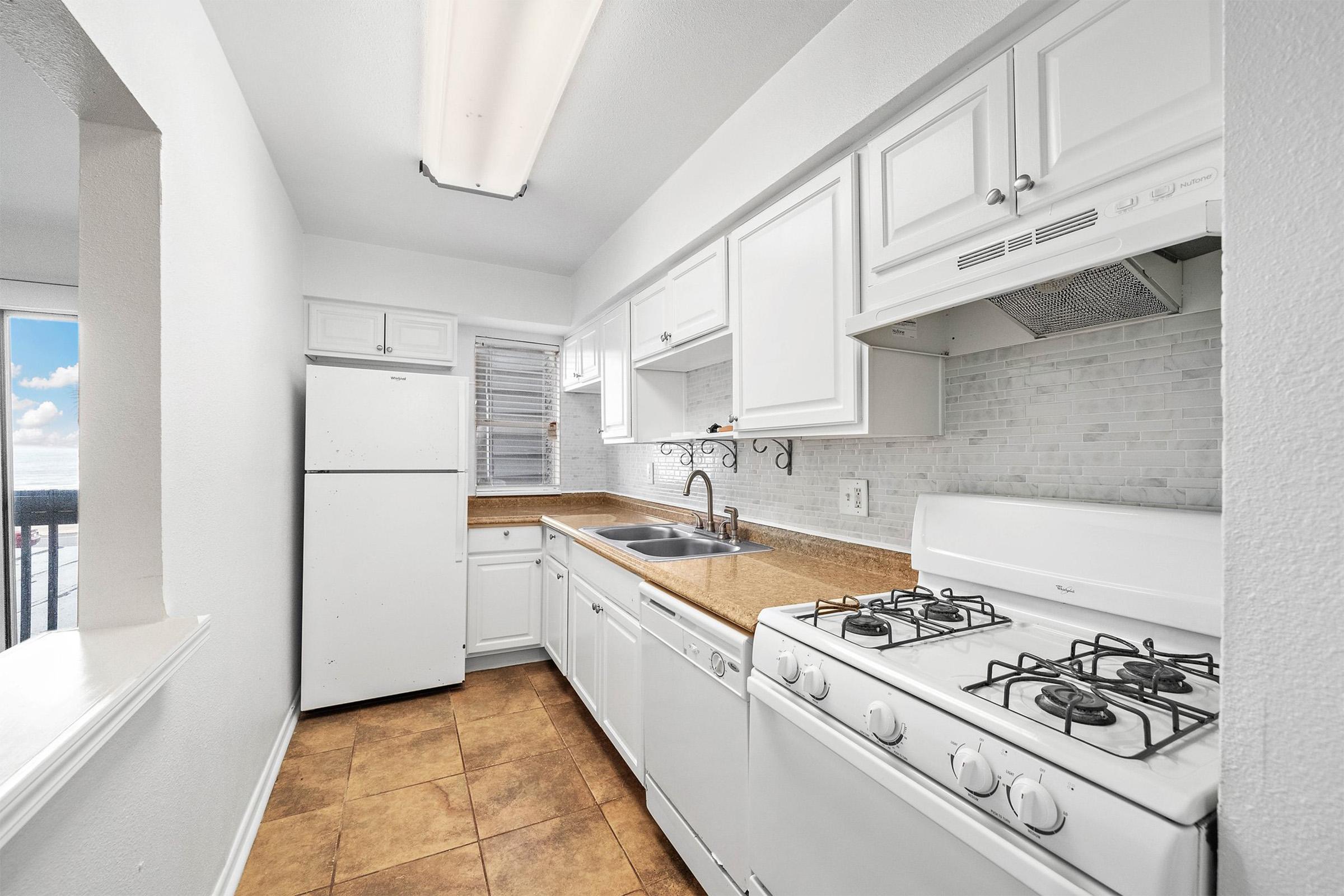 A modern kitchen featuring white cabinetry, a stainless steel sink, a gas stove, and a refrigerator. The counter is made of light-colored material, and the room is well-lit with natural light coming in from a window. Tile flooring adds a stylish touch to the space.