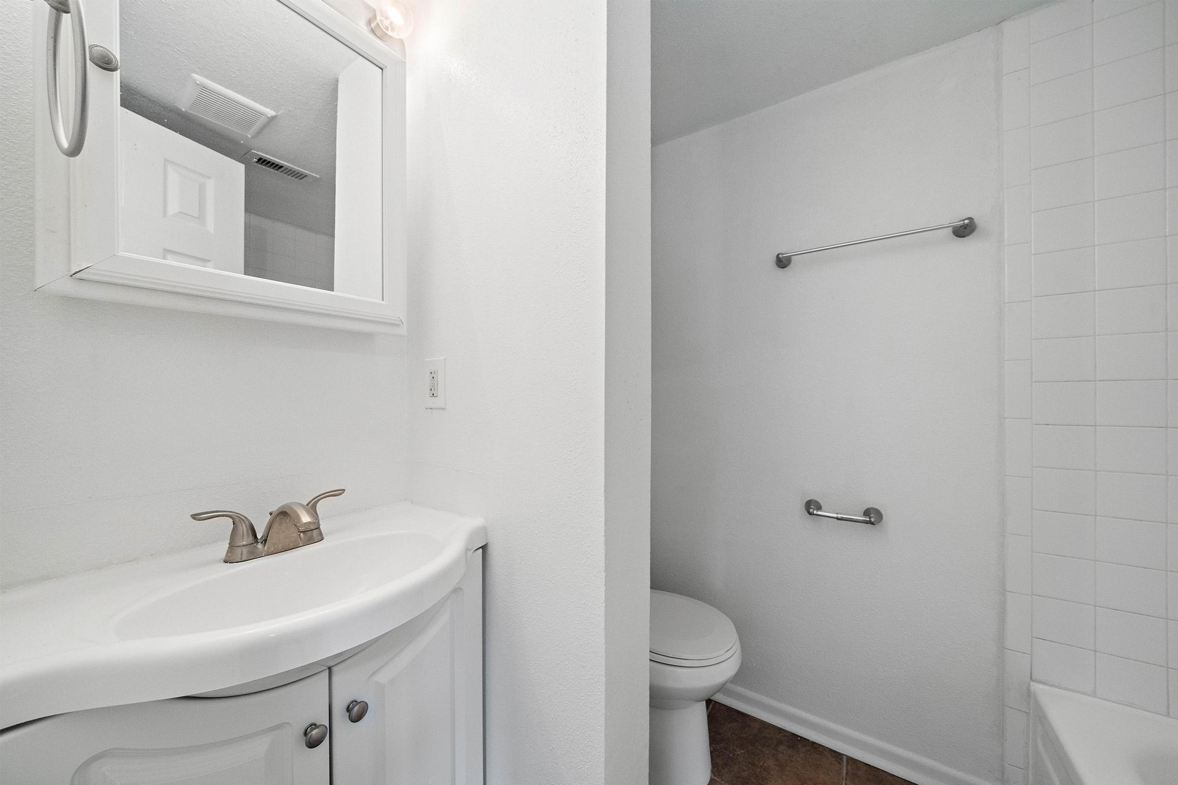 A bright, minimalistic bathroom featuring a white vanity with a sink, a large mirror, and a shower area with tiled walls. The space is well-lit with neutral-colored walls and a simple design. A toilet is visible, and the flooring has a warm, textured look.