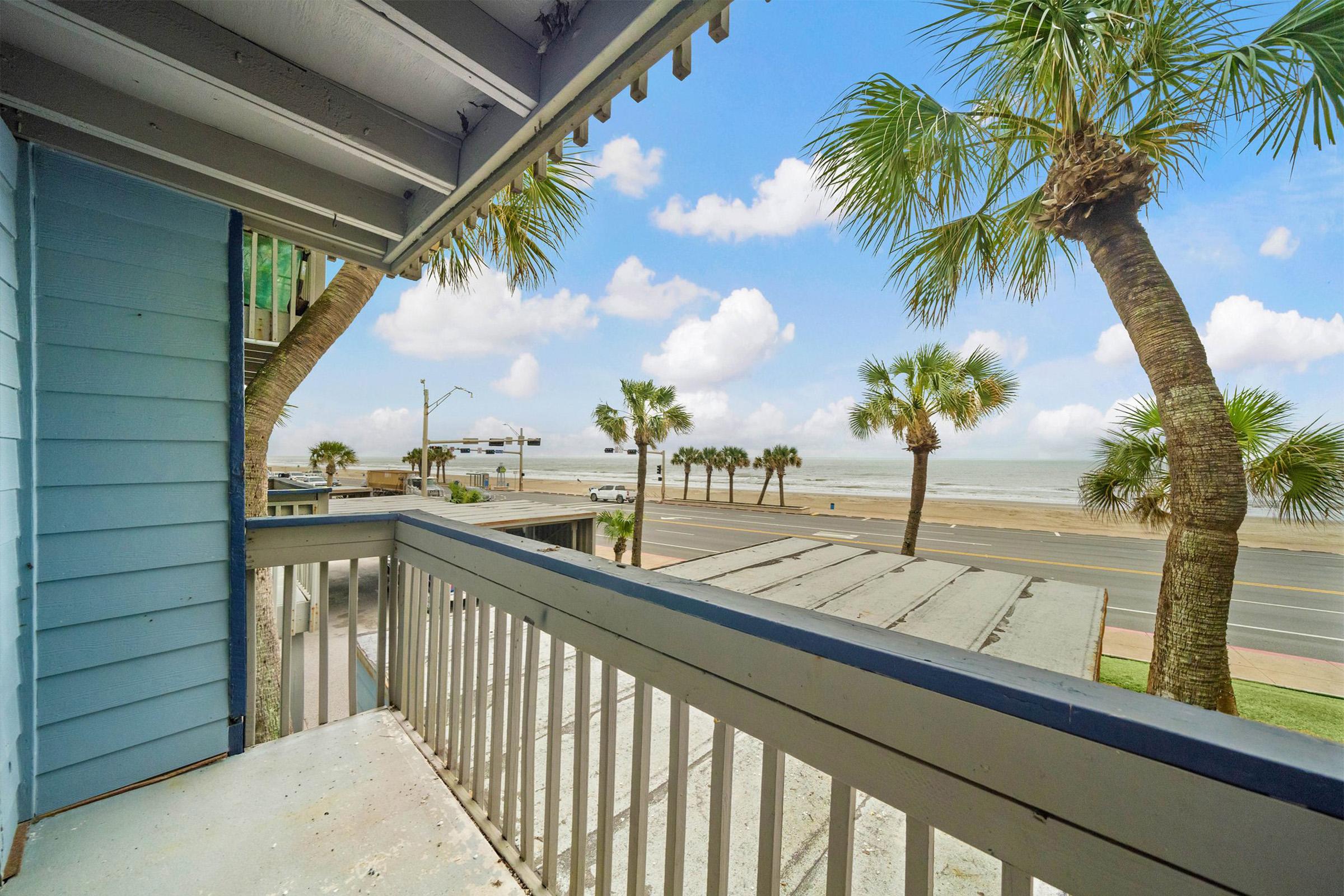 View from a balcony overlooking a road lined with palm trees and a beach. The sky is partly cloudy, and the ocean can be seen in the distance. The scene conveys a tranquil coastal atmosphere.