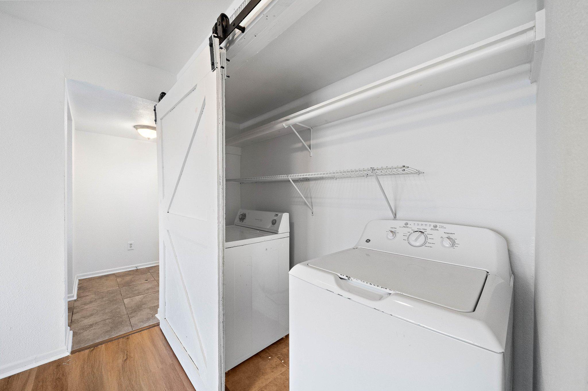 A laundry room featuring a white washer and dryer inside a closet-like space. The area has light-colored walls and a wooden floor, with a metal shelving rack above the appliances. The entrance to the room is visible, along with a bright light fixture in the ceiling.