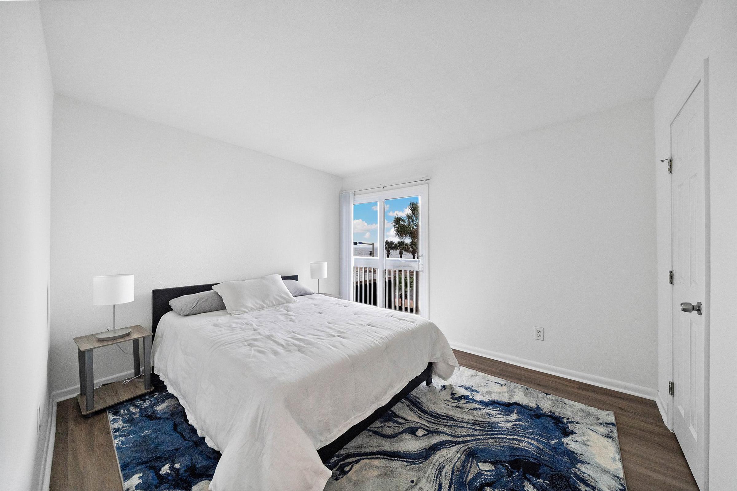 A spacious and modern bedroom featuring a queen-size bed with white bedding, two bedside lamps, and a wooden nightstand. The room has light-colored walls and a large window offering a view of the outdoors, complemented by a blue and white area rug on the wooden floor.