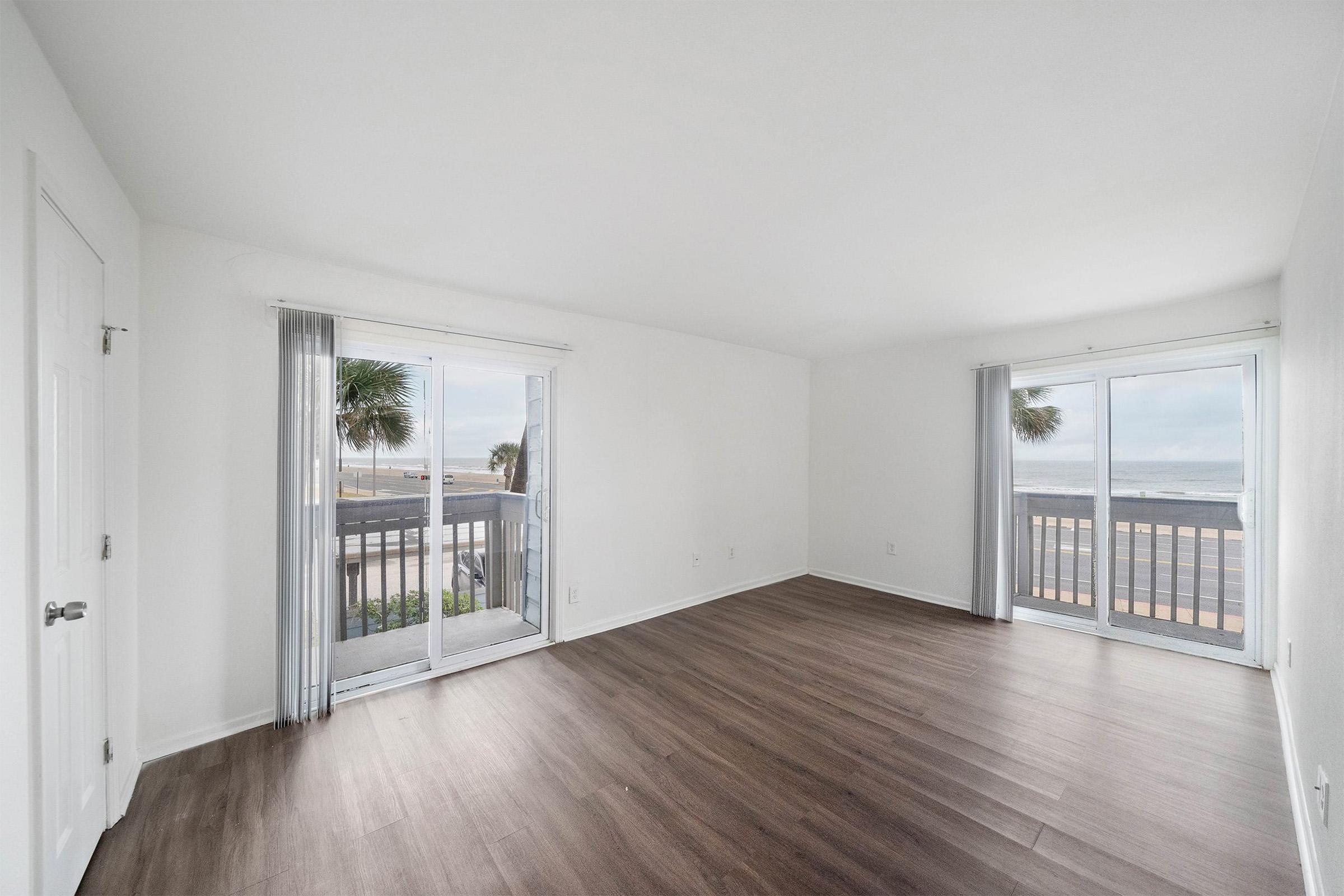 A bright and airy room with light-colored walls and large windows, offering a view of the beach and ocean. The flooring is dark wood, and there are sheer curtains at the windows. The space is empty, creating a clean and minimalist atmosphere.