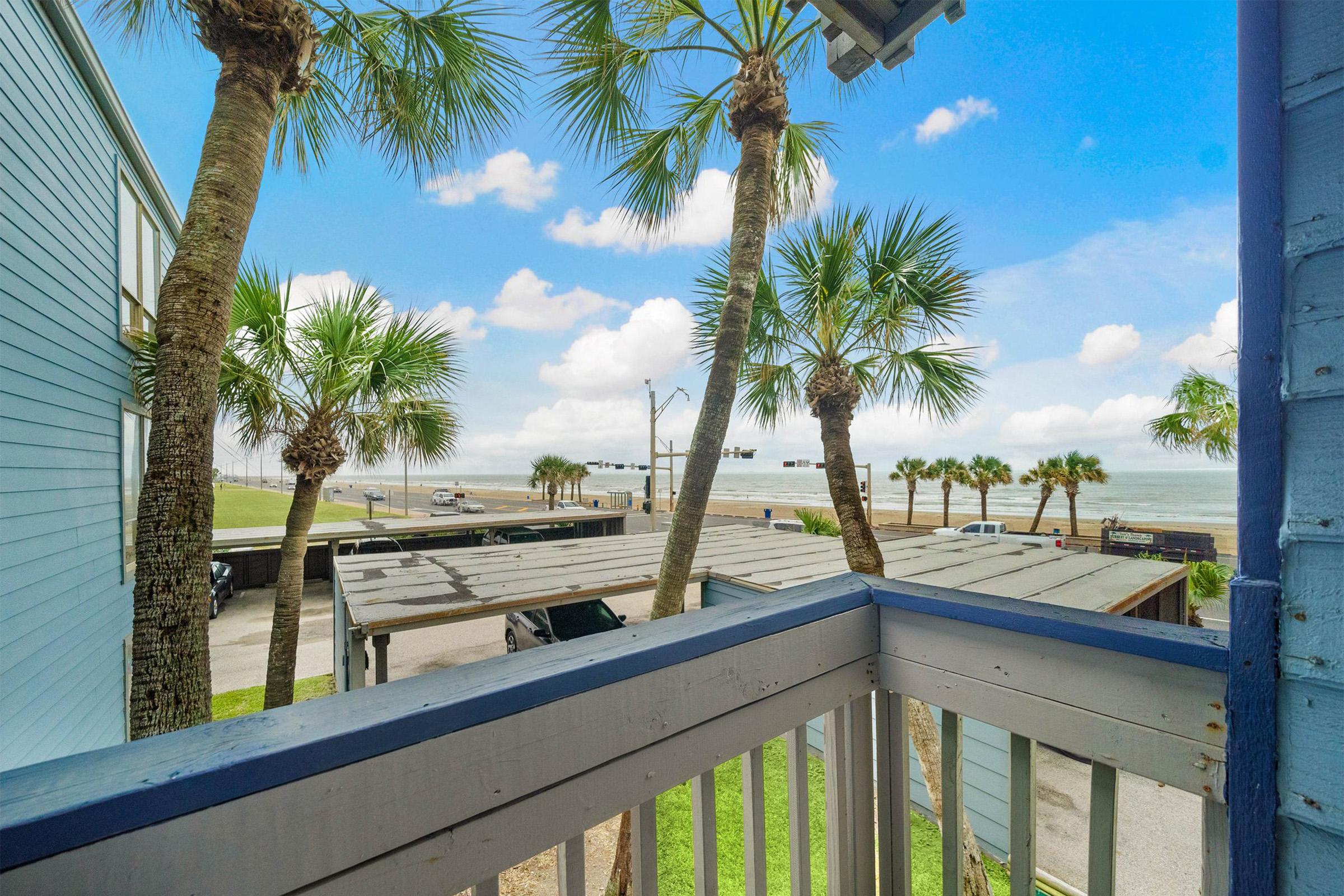 View from a balcony featuring palm trees against a bright blue sky and clouds. In the background, the ocean is visible along with a road and a traffic signal. The scene suggests a peaceful coastal environment.
