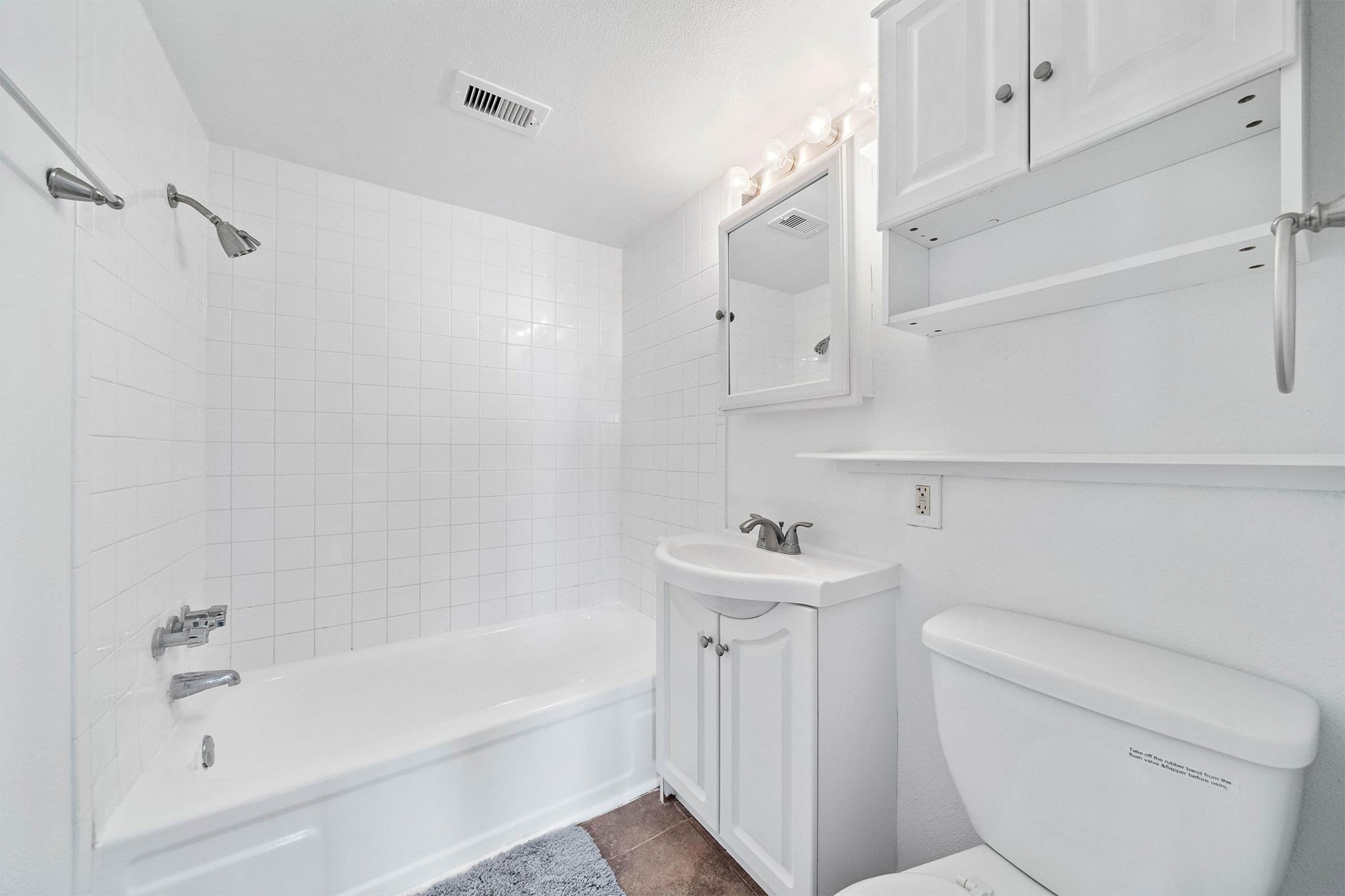 A clean, modern bathroom featuring a white tiled wall, a bathtub with a showerhead, a white vanity with a sink and mirror, and a toilet. Soft lighting is provided by wall-mounted fixtures above the mirror, and there are open shelves for storage. The floor is covered with a neutral-colored rug.