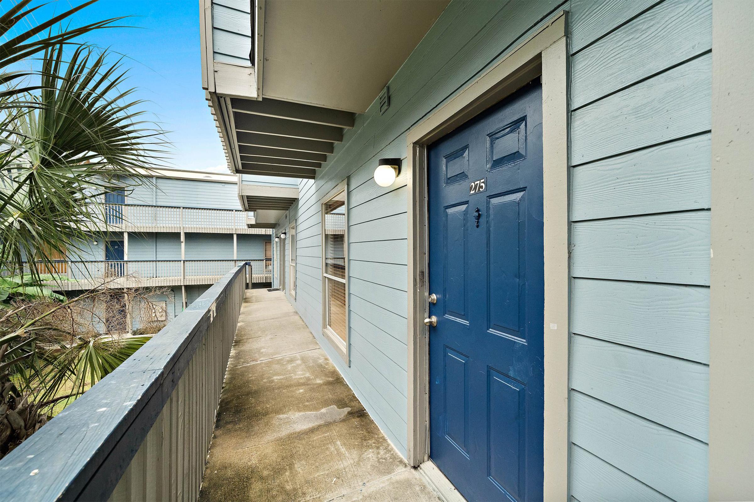 A low-angle view of a blue-painted apartment building corridor featuring a blue door with the number "275" visible. The exterior has light blue wooden siding and a concrete walkway lined with palm trees and other greenery. Bright daylight enhances the scene.