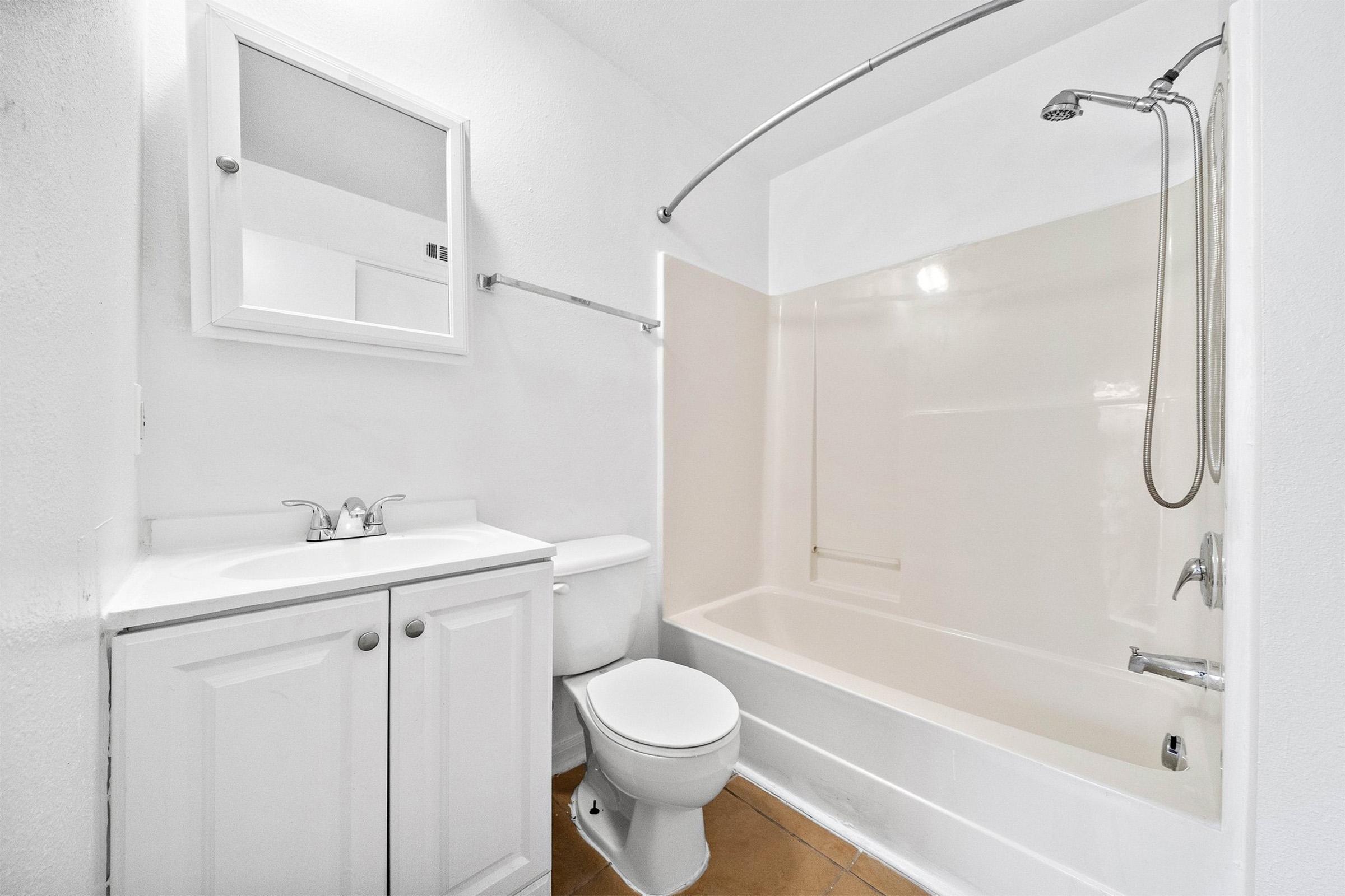 A clean, modern bathroom featuring a white bathtub with a shower, a toilet, and a sink with storage cabinets below. The walls are painted white, and there is a mirror above the sink. The flooring is tiled in a light color, creating a bright and fresh atmosphere.