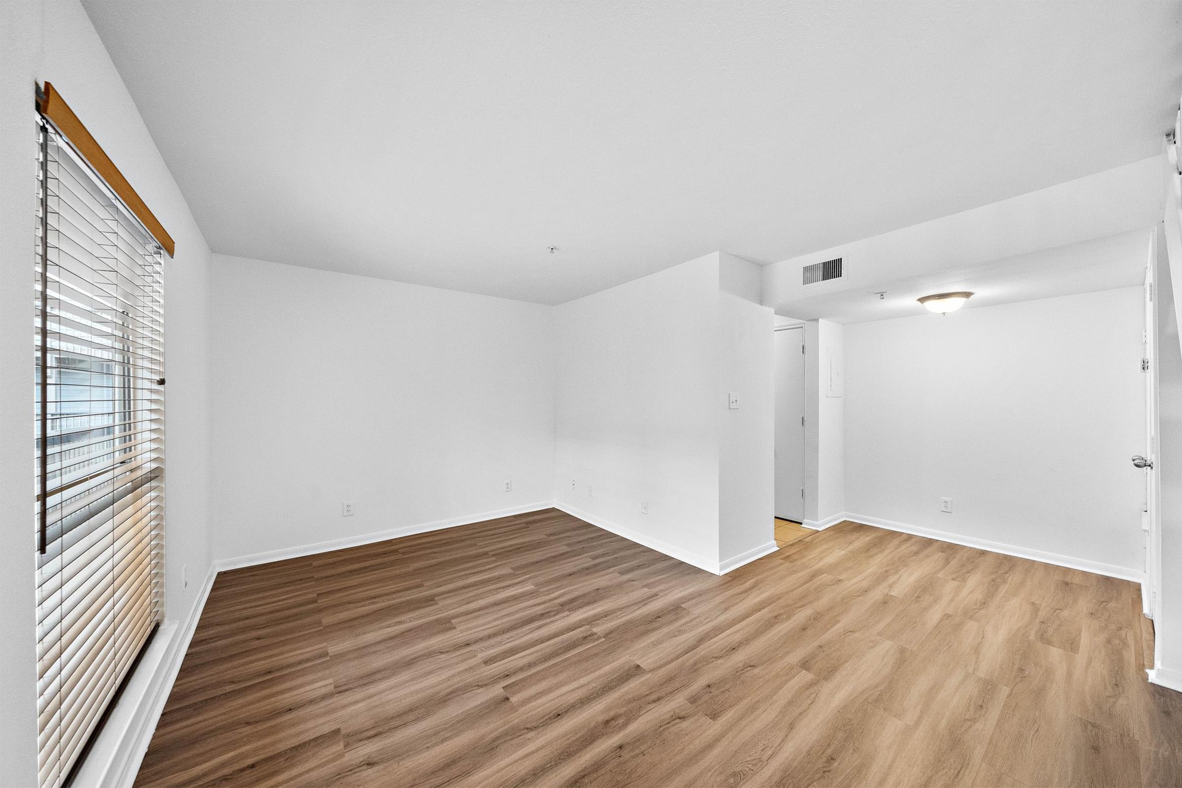 A bright, empty room with light-colored walls and wooden flooring. There are white blinds on a window, and a ceiling light fixture is visible. A doorway leads to another space, suggesting an open layout. The overall atmosphere is clean and spacious, ideal for personalization.