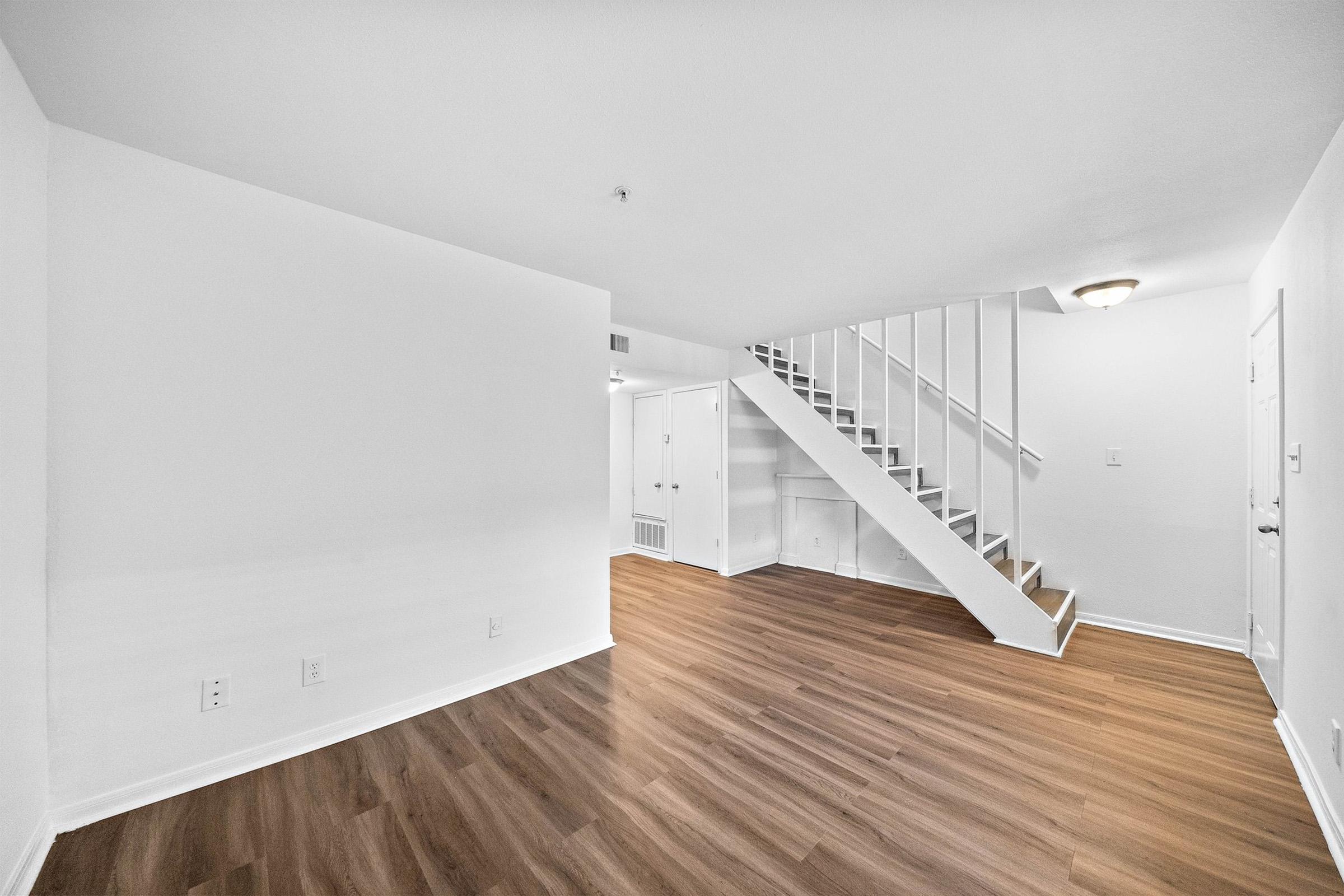 A spacious interior of a residential unit featuring light-colored walls and new hardwood floors. The image shows a staircase leading to an upper level, with a doorway visible on the right. The space is empty, creating a bright and open atmosphere.