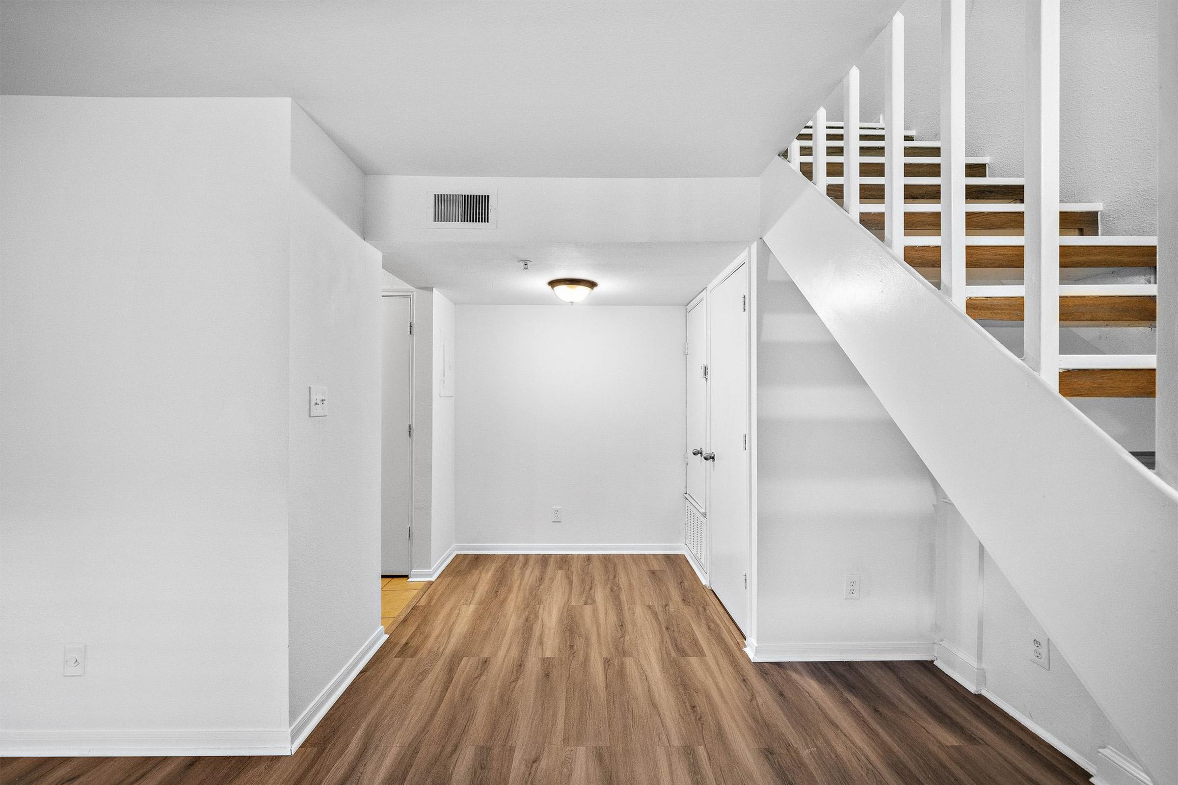 A well-lit interior of a home featuring an open space with wooden flooring. A staircase with white railings is visible on the right side, leading to an upper level. The walls are painted white, and there are doors to the left, indicating entrance points to other areas.