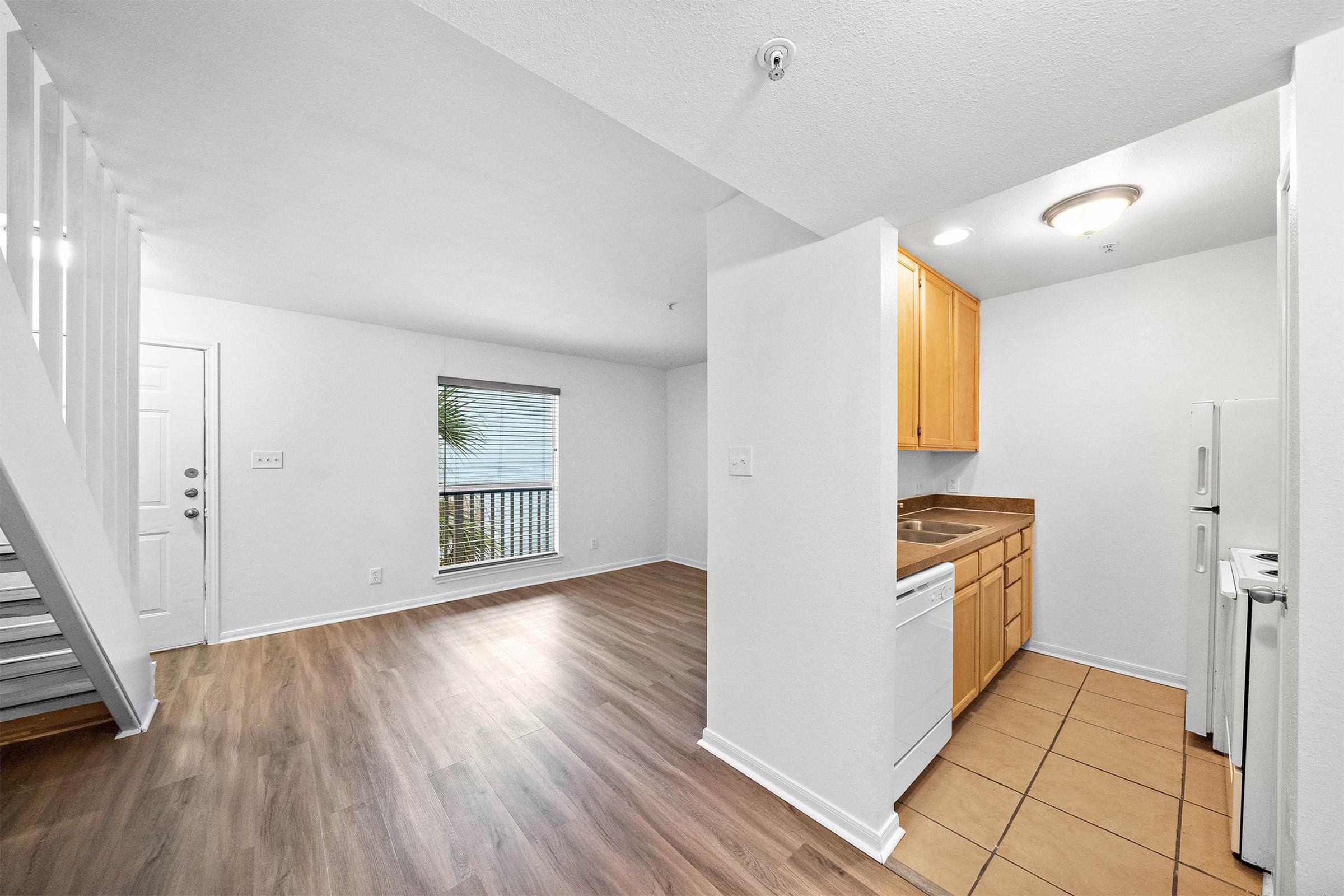 A bright and spacious living area with light-colored walls and wood-style flooring. On the left, there’s a set of stairs leading to the upper level. The kitchen features wooden cabinets, a sink, and an adjacent refrigerator. A large window lets in natural light, offering a view of a balcony.