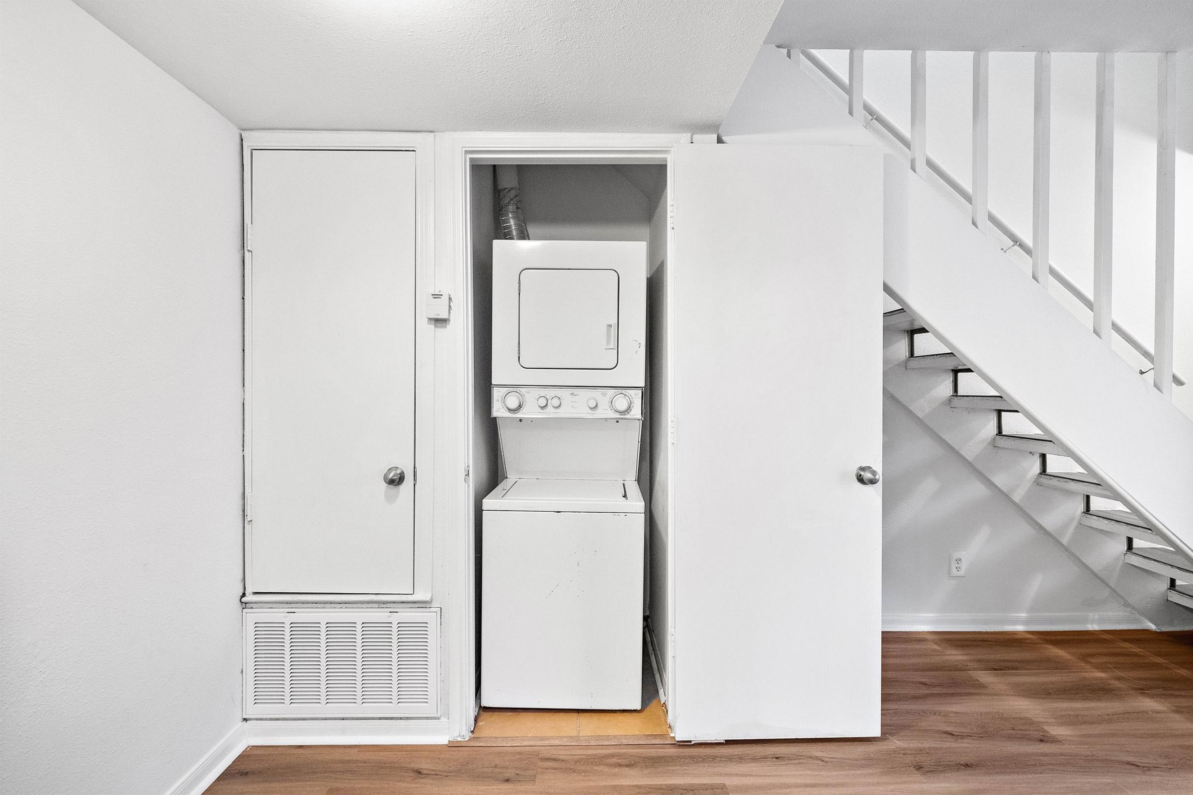 A small laundry area featuring a stacked washer and dryer unit tucked into a closet space. The closet doors are open, revealing the appliances. Light-colored walls and wooden flooring create a bright and clean environment, with a staircase visible in the background.