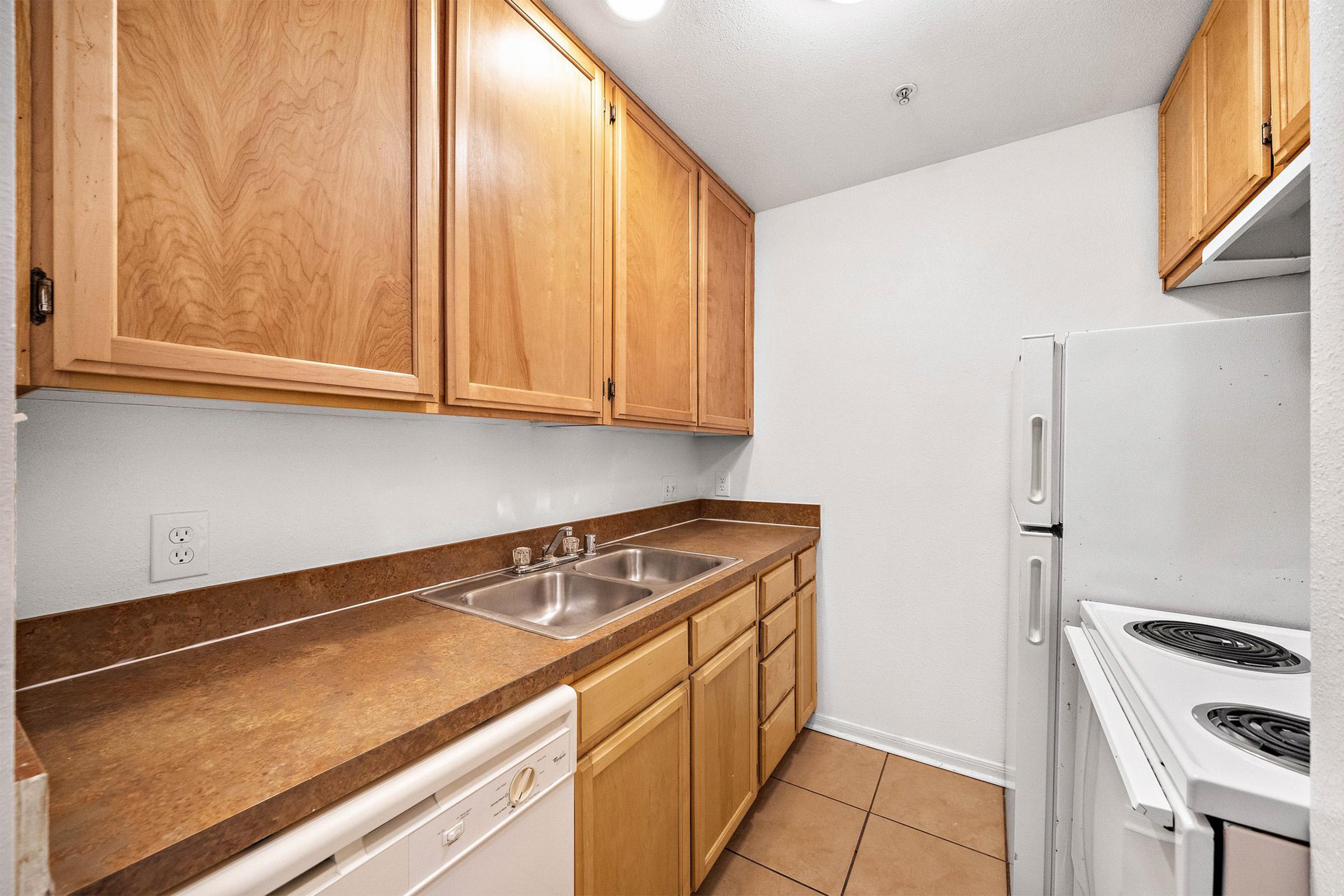 A small kitchen featuring wooden cabinets, a double sink, a white dishwasher, and a stove with an oven. The countertops are a brown laminate, and the floor is tiled. The walls are painted white, creating a bright and simple cooking space.