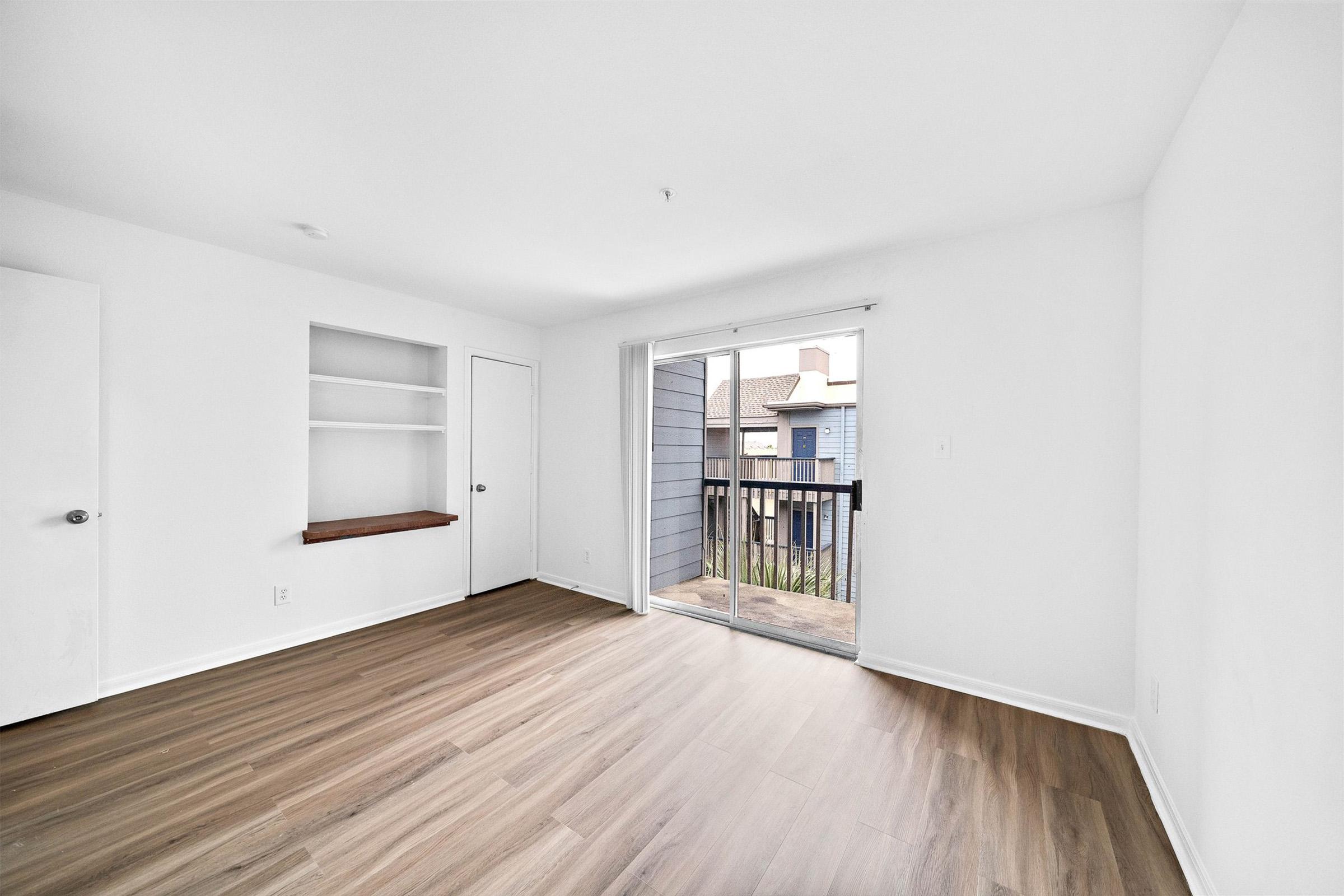 A bright, empty room with white walls and wooden flooring. There is a large sliding glass door leading to a small balcony, allowing natural light to enter. A built-in shelf is positioned against one wall, and a closed door is visible on the left side of the image. The space is unadorned and ready for furnishing.