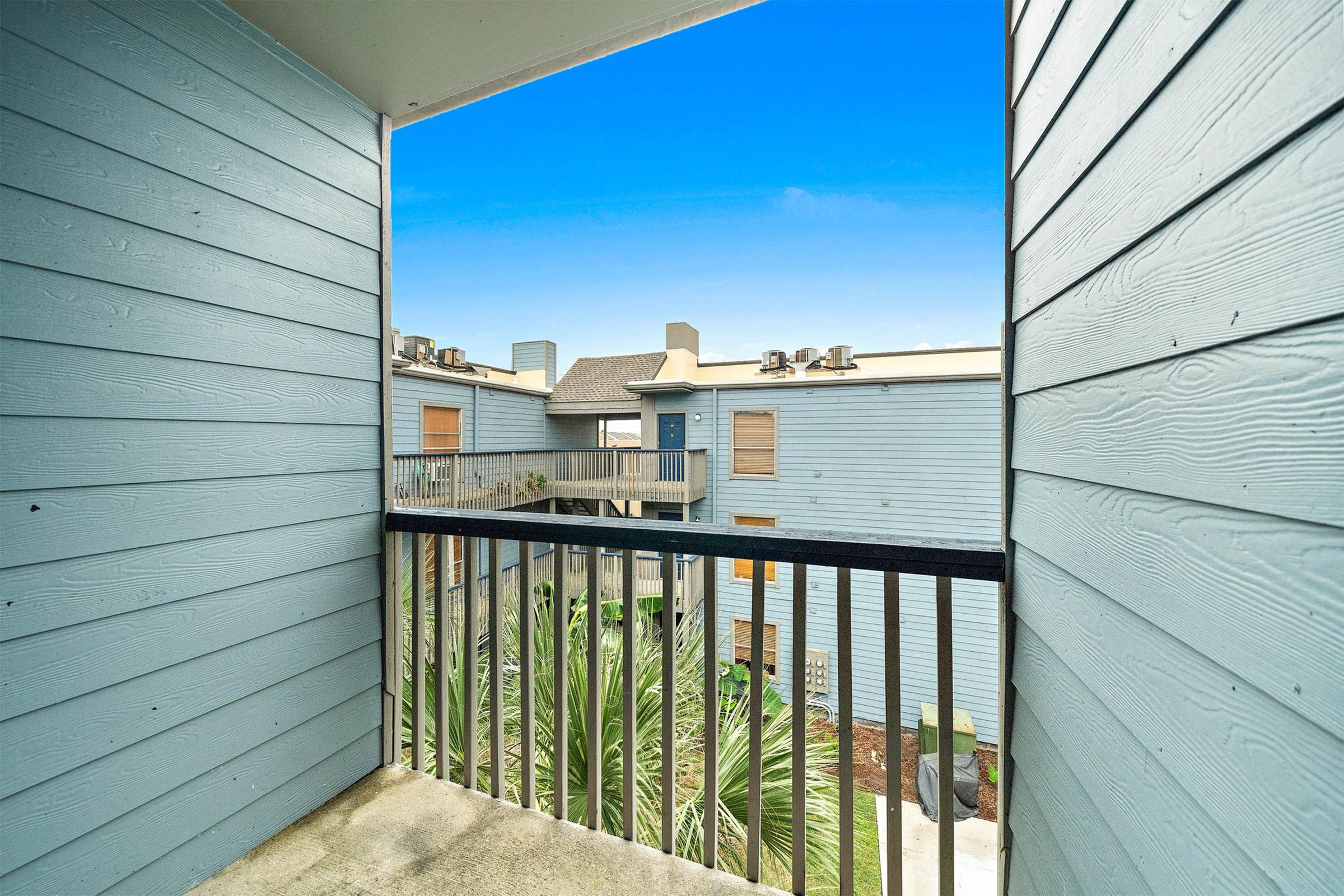 View from a balcony overlooking an apartment complex. The scene features light blue wooden siding, a clear blue sky, and a glimpse of palm trees and landscaped areas in the courtyard. The setting has a peaceful, residential atmosphere.