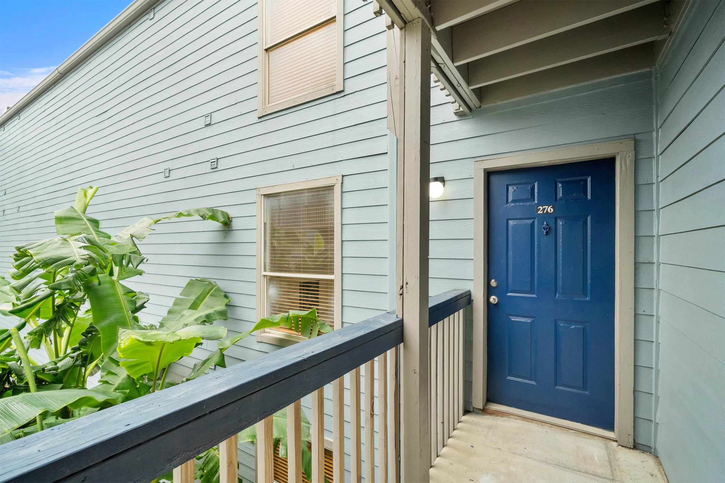 A blue front door with the number 276 is situated on a light blue exterior wall. The entrance is framed by a wooden railing, and lush green plants are visible nearby. Natural light filters in through nearby windows.