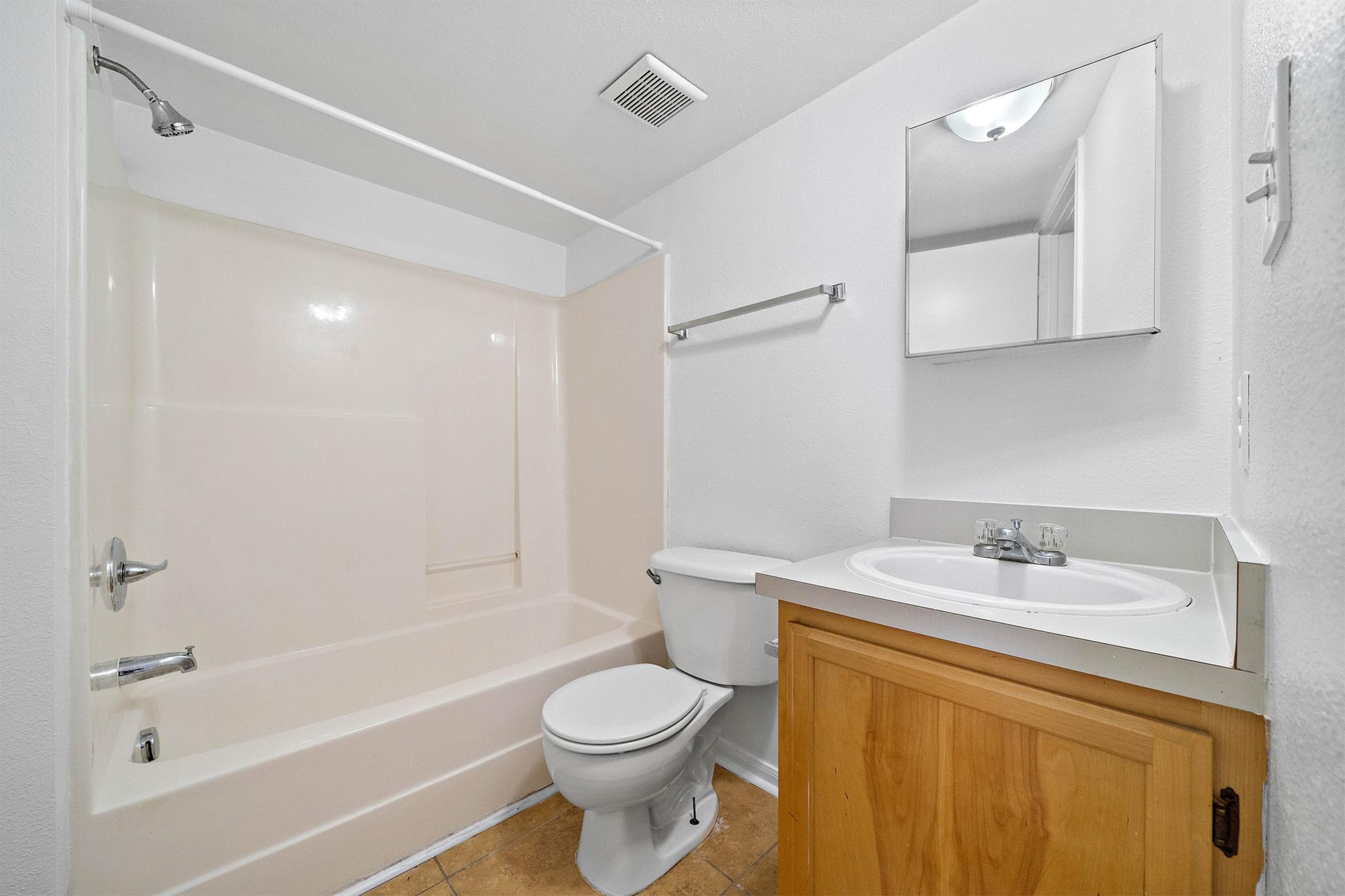 A small bathroom featuring a bathtub with a showerhead, a white toilet, a wooden cabinet under the sink, and a mirror above the sink. The walls and ceiling are painted white, and the floor is tiled. The overall appearance is clean and functional.