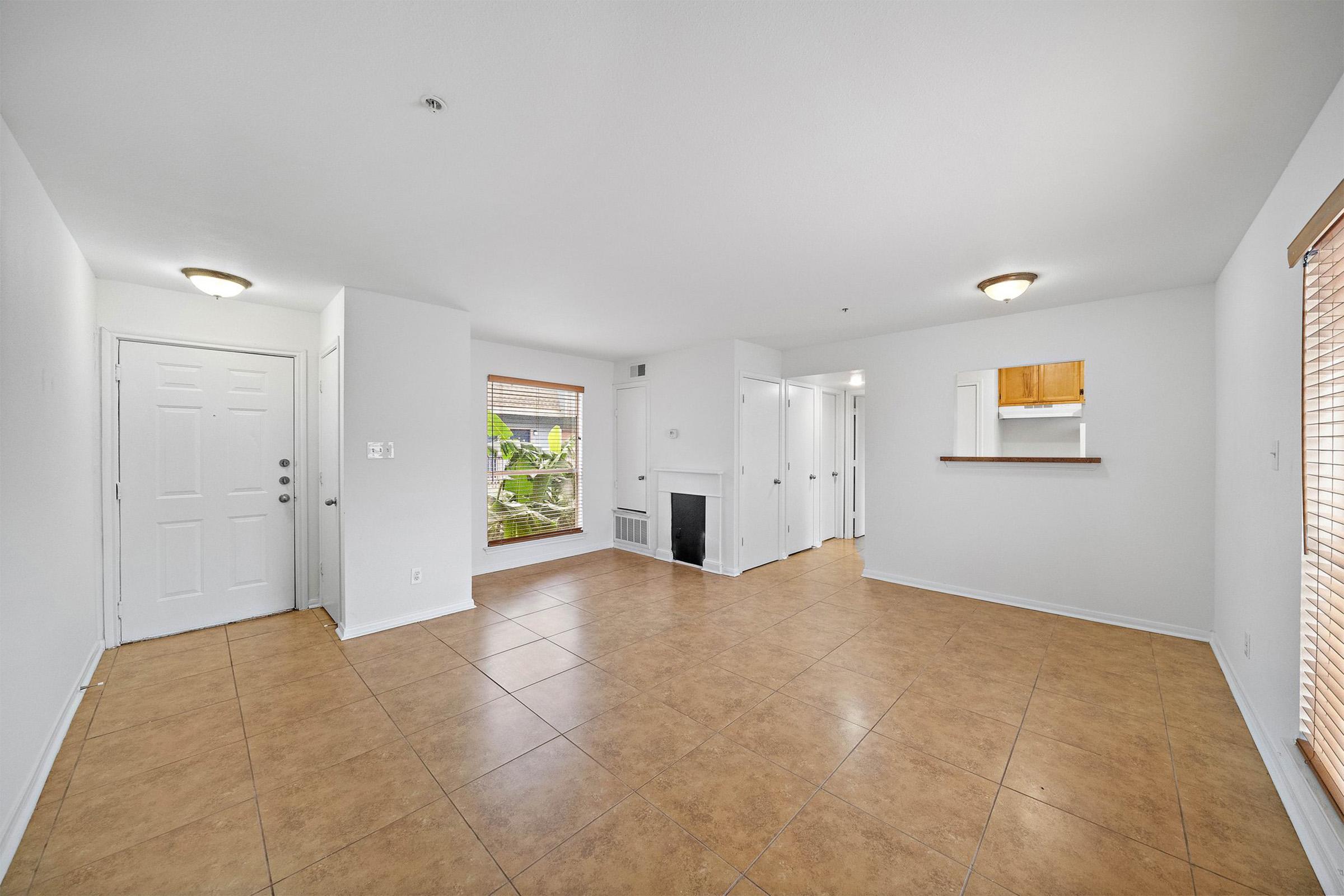 Spacious, bright living area featuring tile flooring, white walls, and natural light from large windows. A small adjacent kitchen area is visible, with a bar counter separating it from the living space. The room includes a fireplace and a door leading outside, creating a welcoming atmosphere.