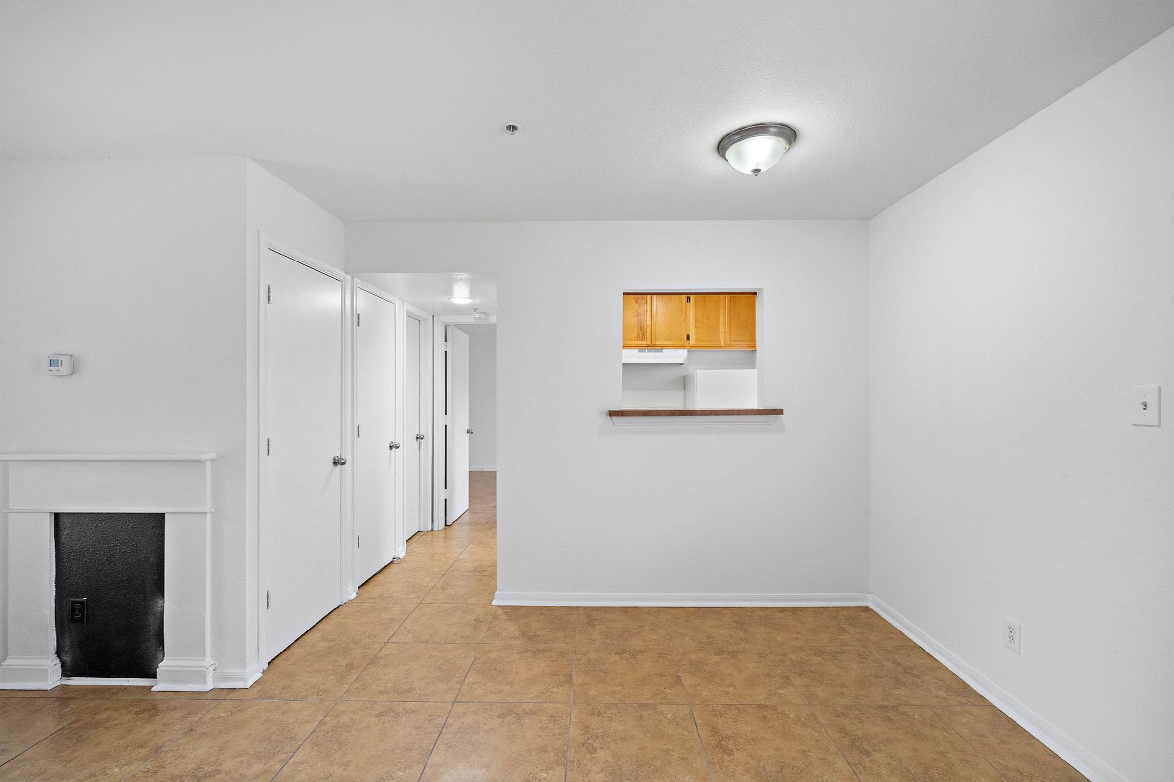 A spacious, empty room with light-colored walls and tile flooring. There is a small shelf opening leading to a kitchen area, a fireplace in one corner, and several doorways leading to other rooms. The ceiling features a light fixture, and the space is filled with natural light.