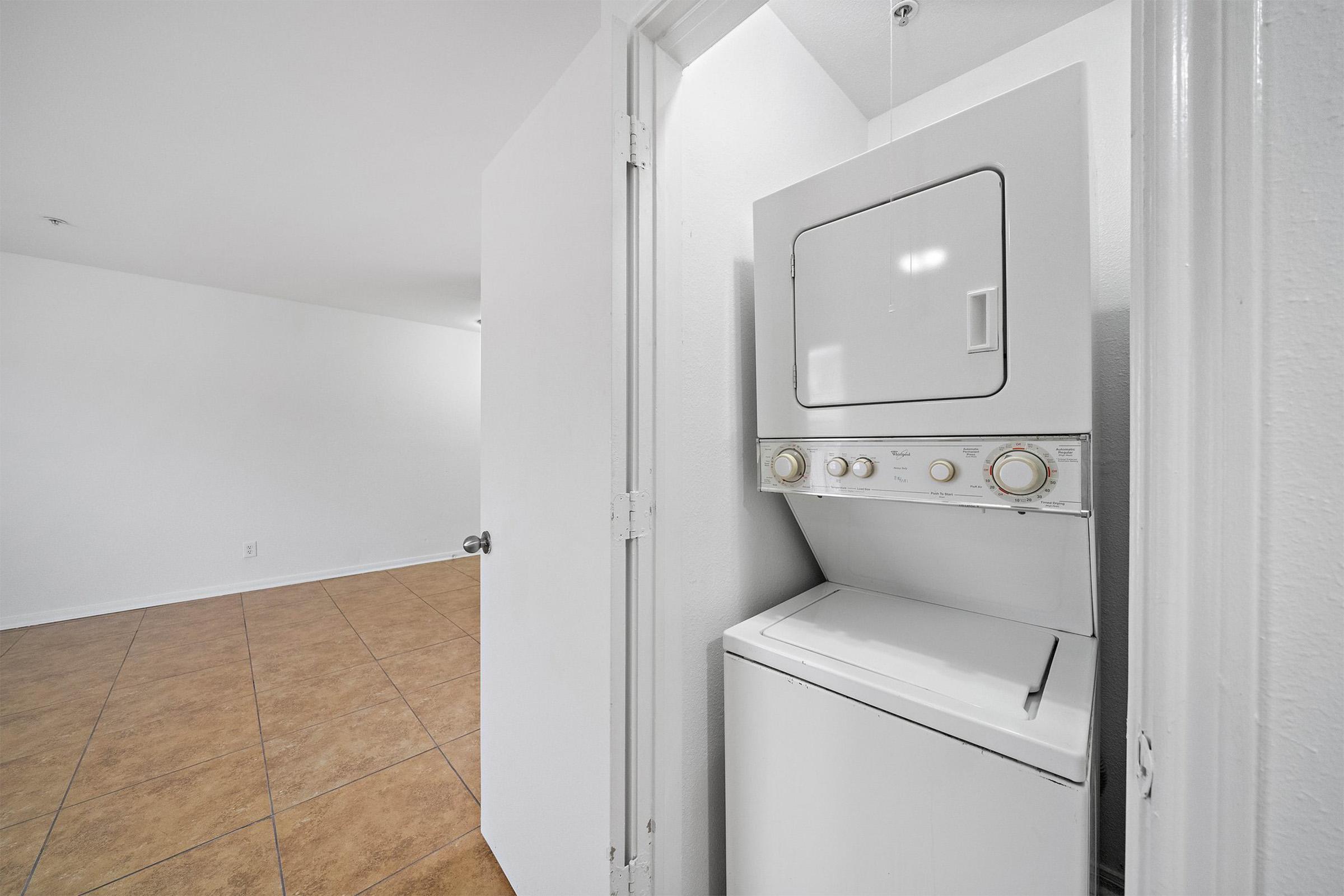 A compact laundry area featuring a stacked washer and dryer unit. The appliance is situated in a corner, with a doorway leading to a spacious, empty room with tiled flooring and light-colored walls.