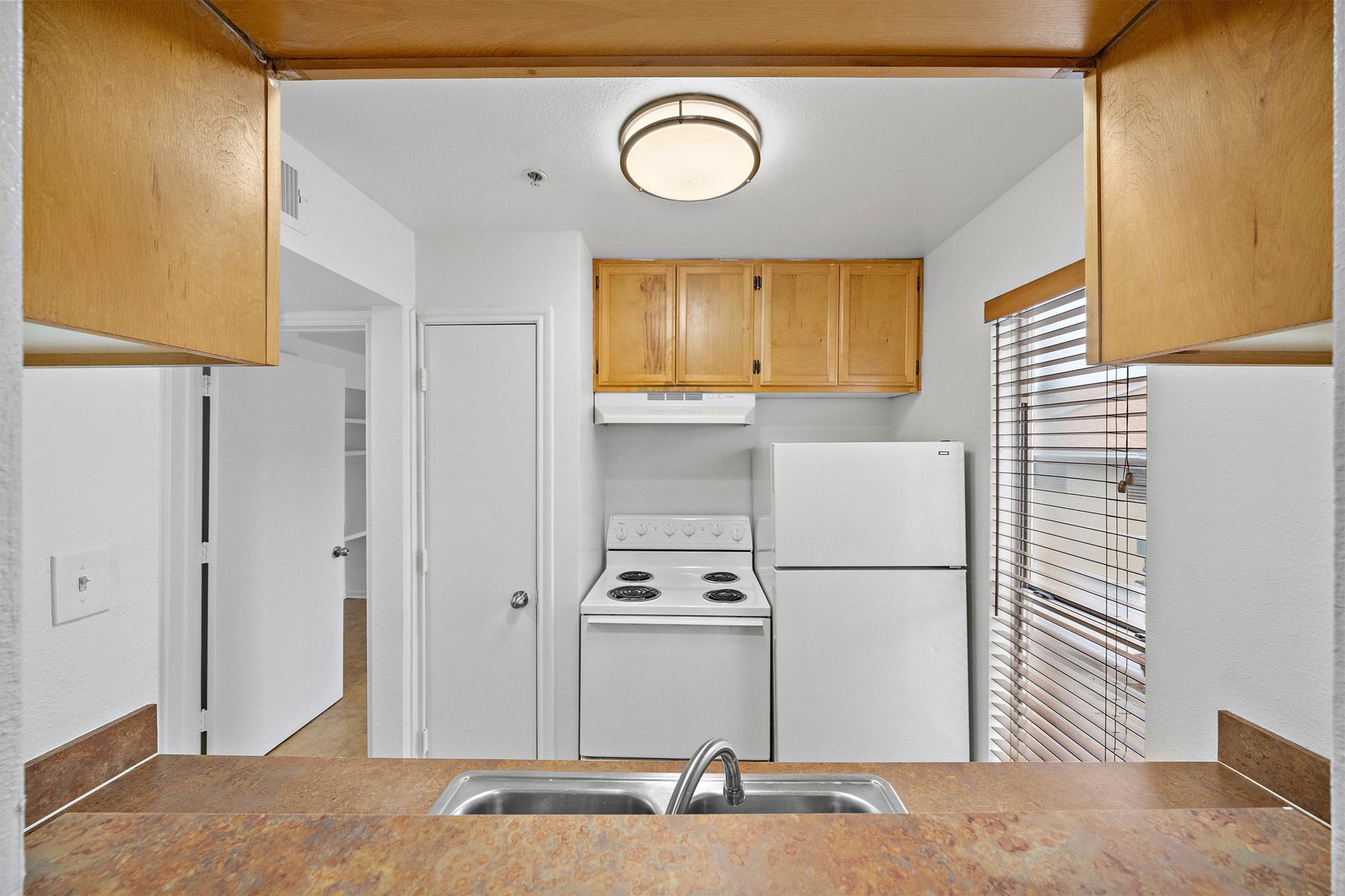 A kitchen view featuring a sink in the foreground, with a white stove and refrigerator visible. The cabinets above are made of light wood, and there is a window with blinds letting in natural light. The walls are painted white, creating a clean and bright atmosphere.