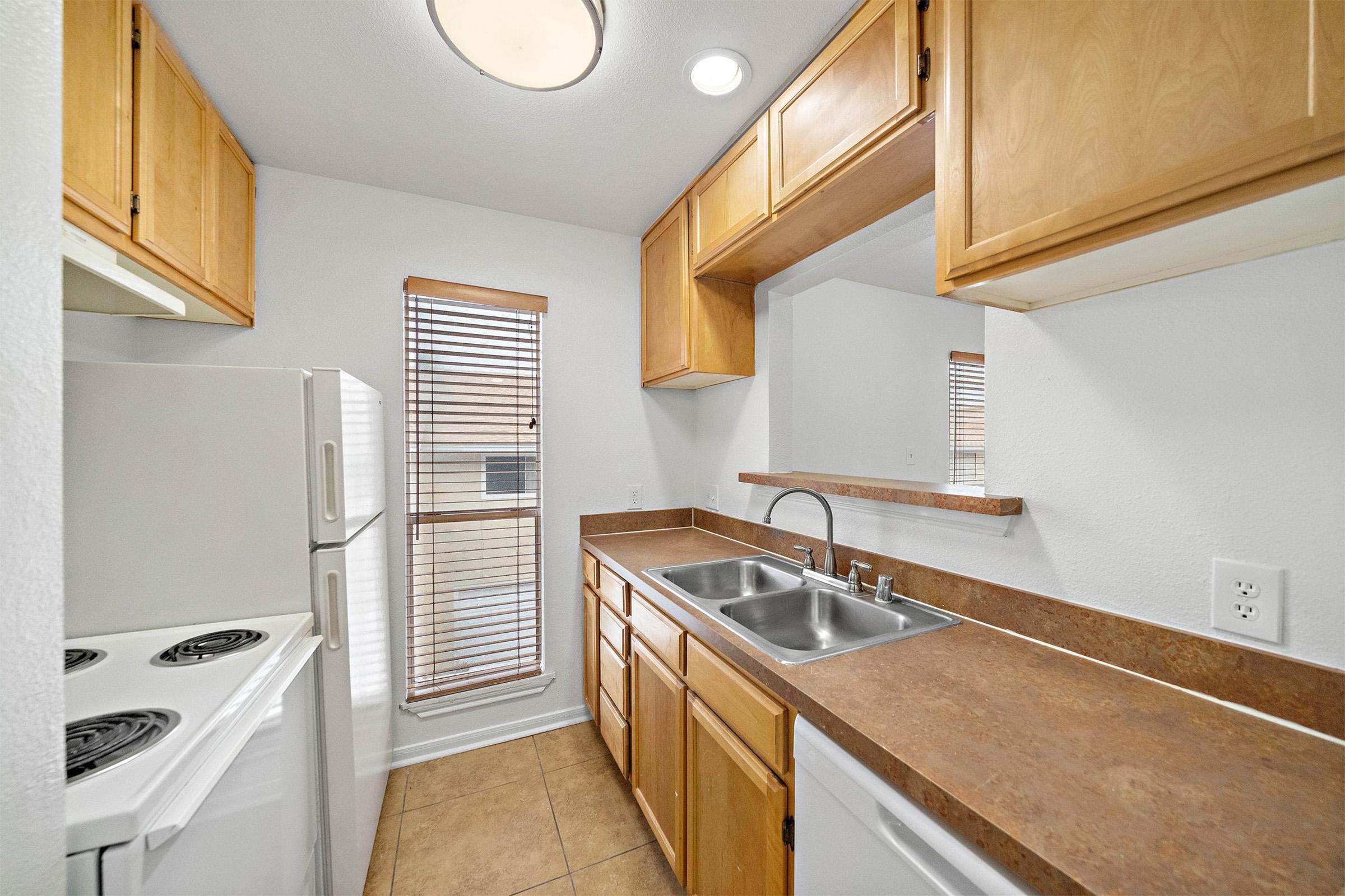 A small kitchen with wooden cabinets, a double sink, and a countertop. The kitchen features a white refrigerator and stove. It has a window with blinds, allowing natural light to enter. The walls are painted white, and the floor is tiled.