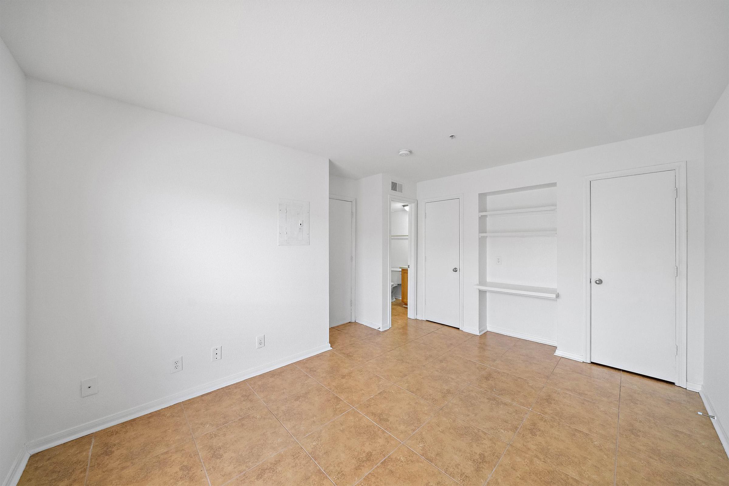 A spacious, empty room with light-colored walls and tan tile flooring. There are two doors on the right side leading to other areas. A built-in shelf is on the left wall, and the overall lighting is bright and neutral, creating an open and airy feel.