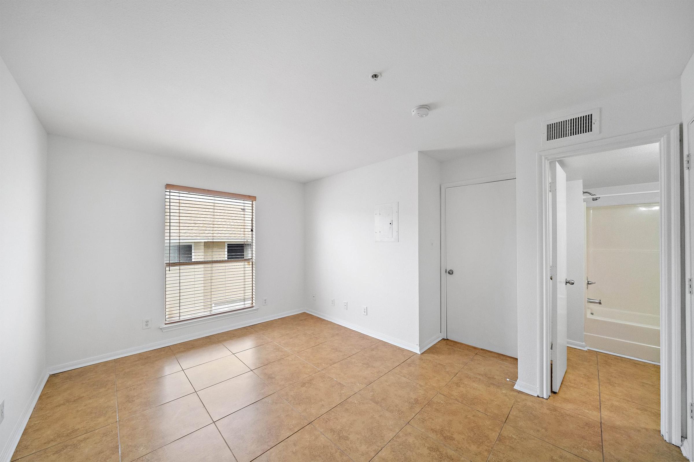 A bright, empty room featuring white walls, a window with blinds, and tiled flooring. There's a door leading to a bathroom visible in the corner, and a small section of the wall is bare, suggesting it may be ready for painting or decoration. Natural light illuminates the space.