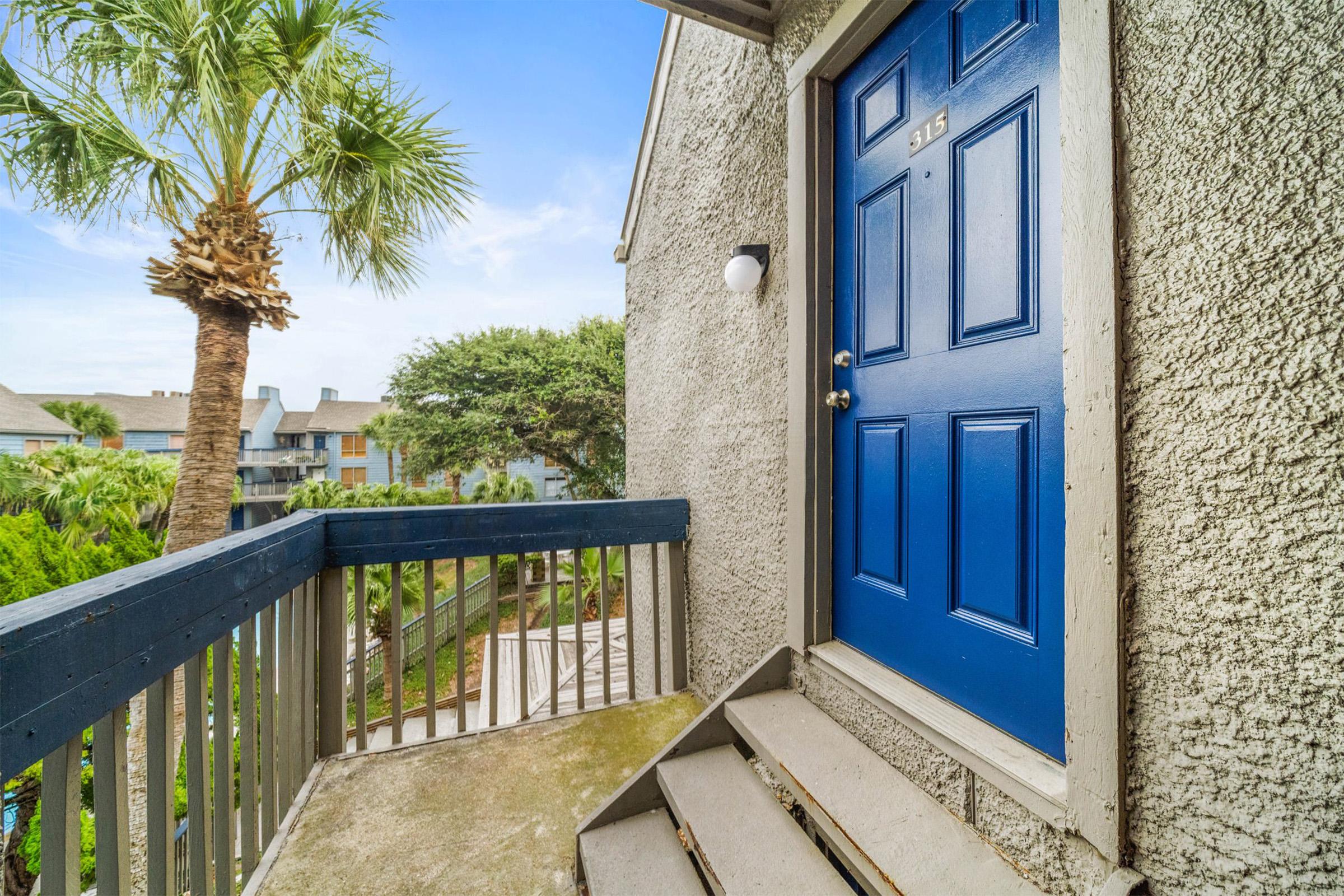 A close-up view of a blue front door on an apartment balcony, with a staircase leading down. In the background, there are palm trees and landscaped greenery, suggesting a tropical or resort-like setting. The sky is clear and bright, contributing to a cheerful atmosphere.