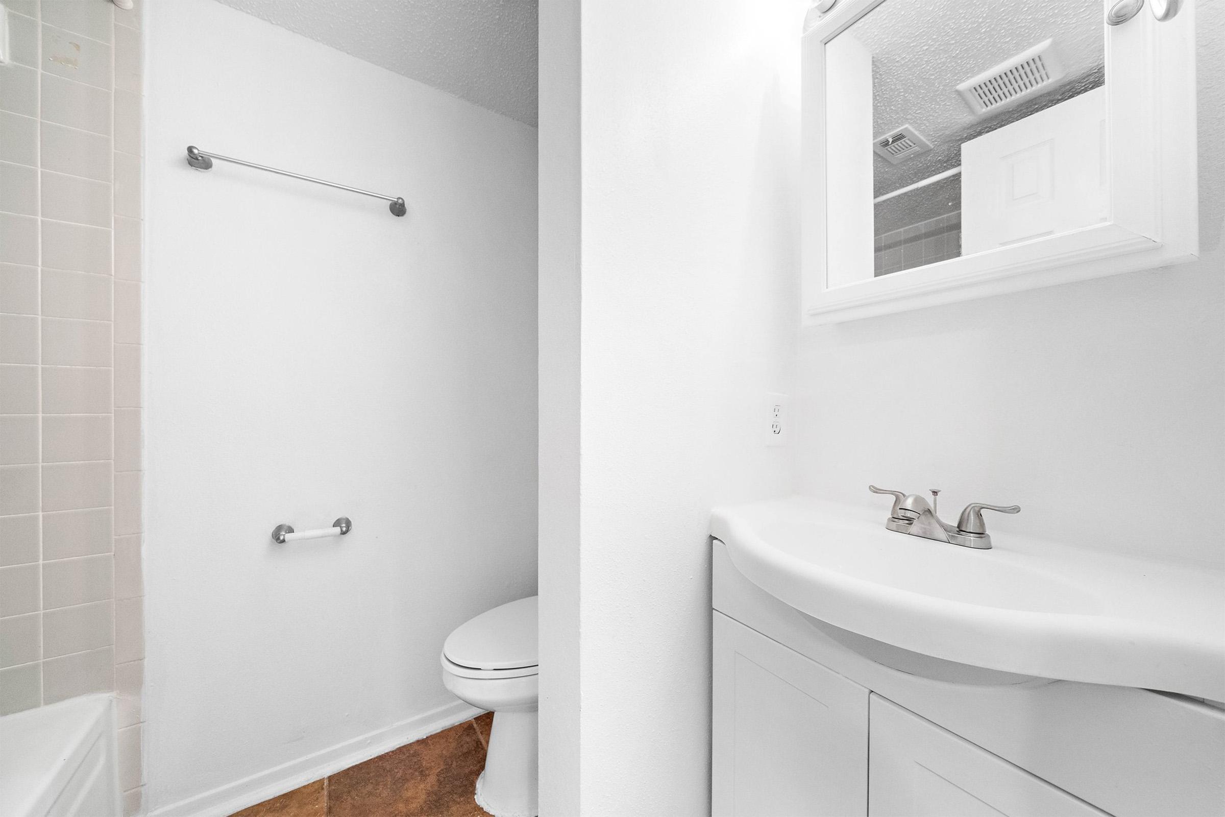 A clean and well-lit bathroom featuring a white sink with two faucets, a toilet beside it, and a bathtub partially visible to the left. The walls and fixtures are mostly white, creating a bright and spacious feel. A mirror above the sink adds to the open ambiance.