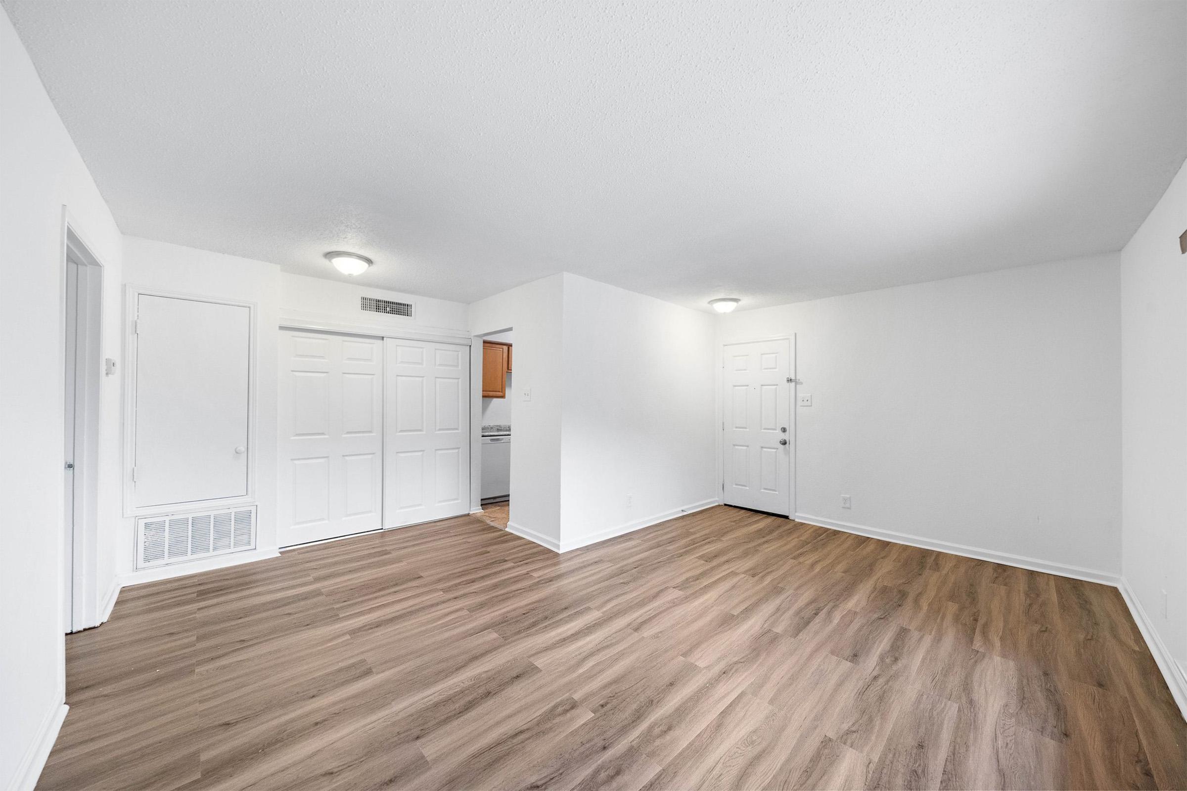 An empty, well-lit living room featuring light wood flooring and white walls. There are two doors leading to other rooms, and a closet with double doors to the left. The space is clean and minimalistic, perfect for furniture arrangement.
